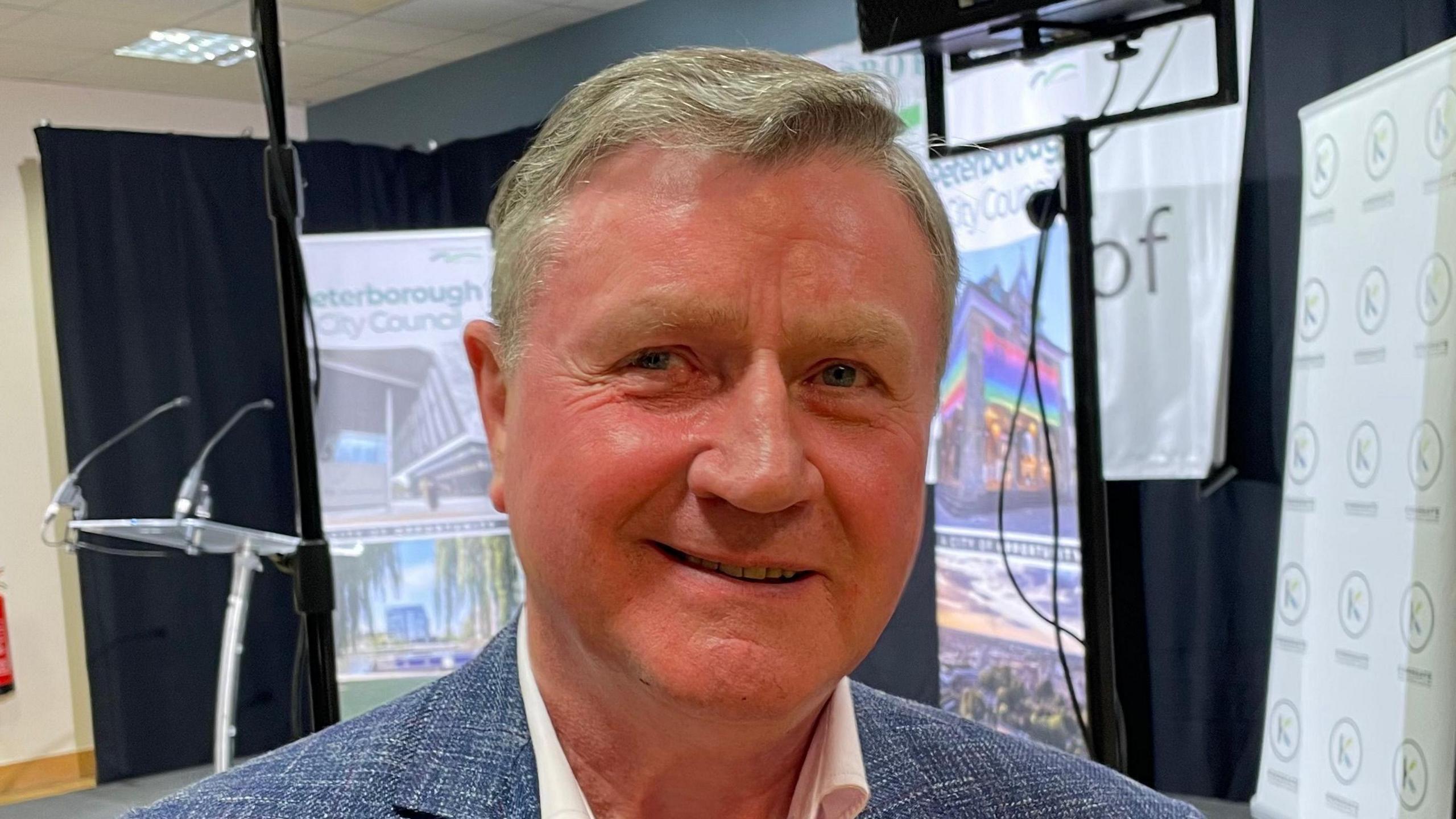 Dennis Jones is wearing a light blue suit and looking at the camera, behind him are some signs with Peterborough City Council written on them