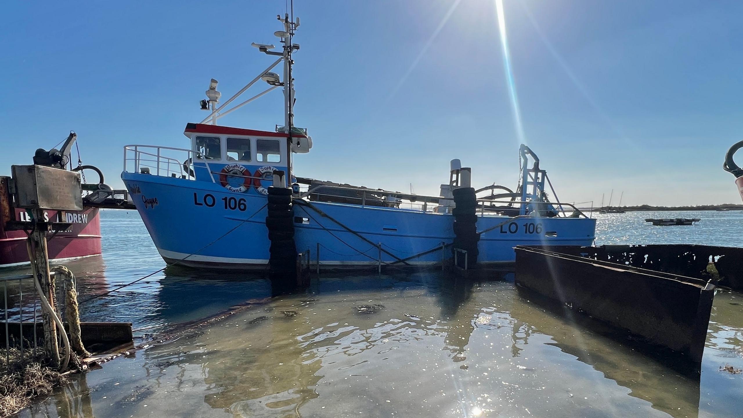 A blue boat with a tall mast sits in a body of water. The water is shining and the sky above is blue with some lens flare.