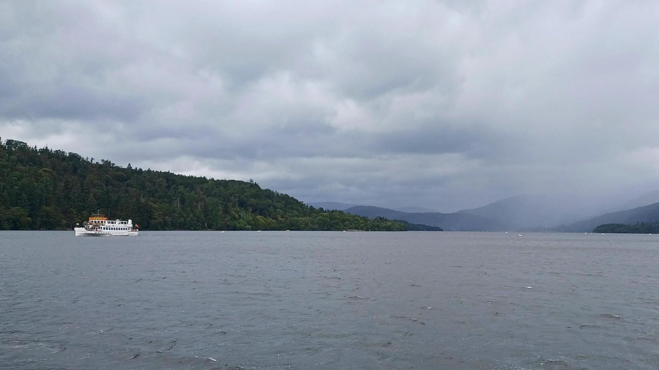 Heavy cloud and rain falling over Windermere as a tripper boat moves through the waters, and small waves are lapping