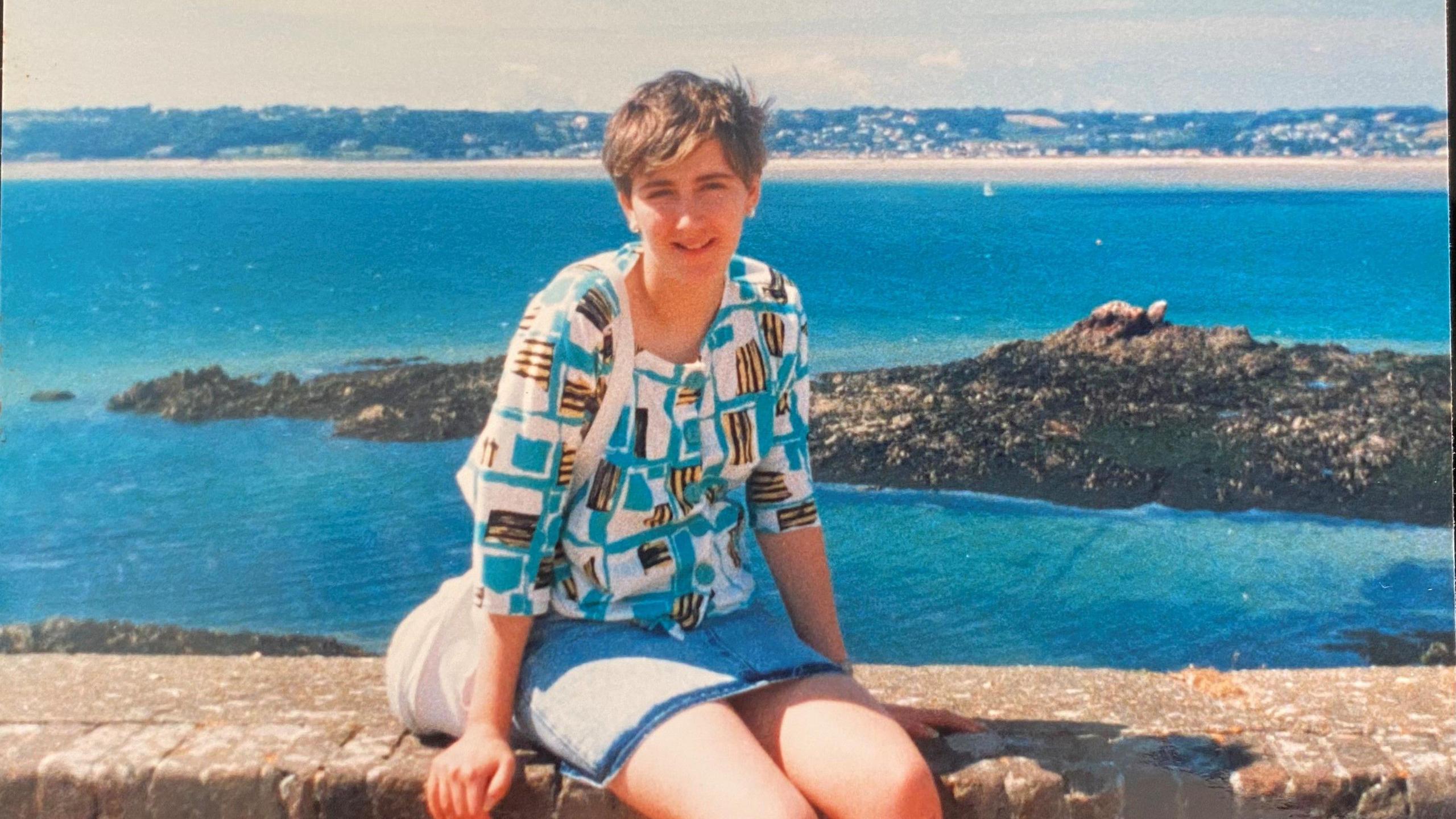 Tina sits on a stone wall with the Jersey sea behind her on a holiday