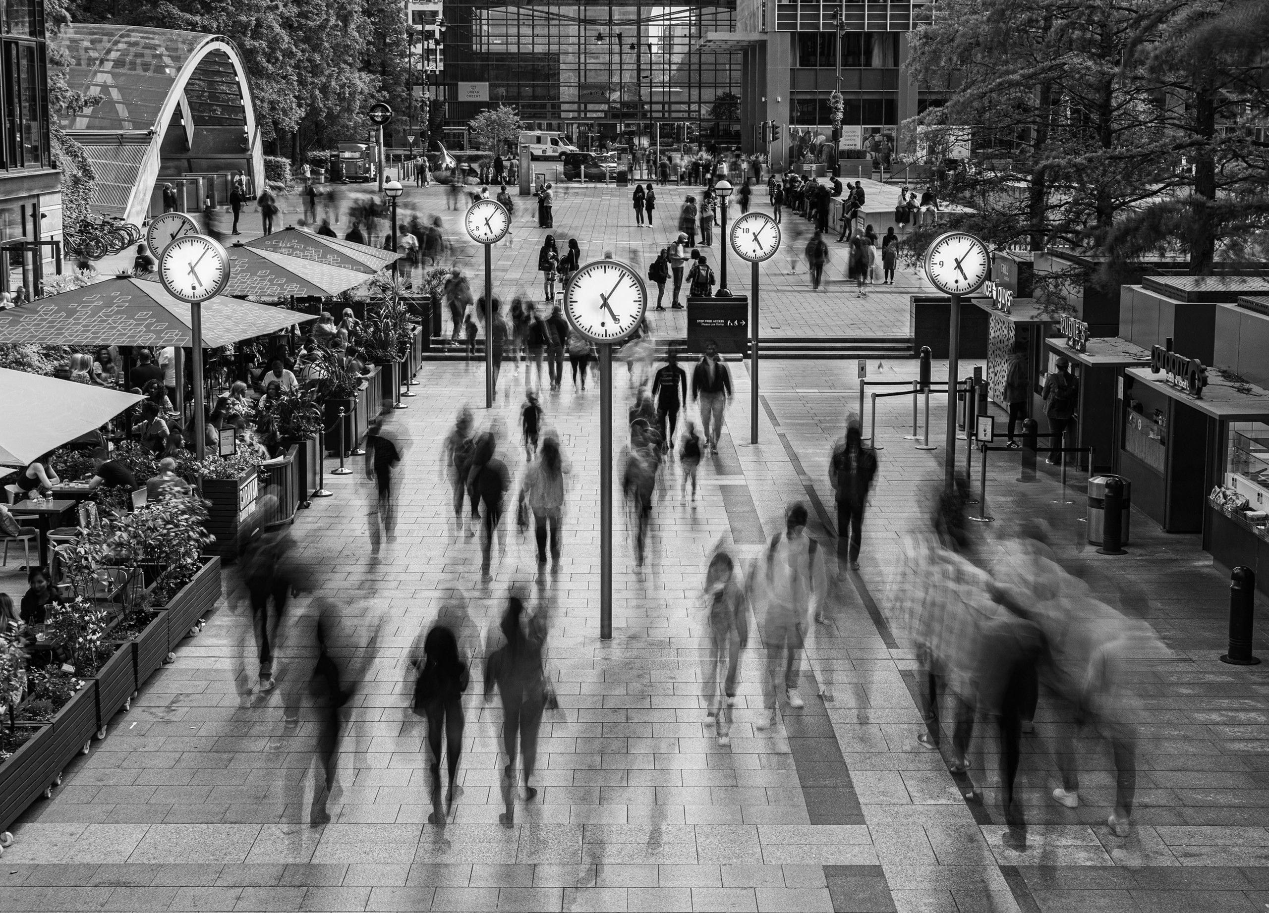 People appear blurred as they walk past five clocks which stand at the top of tall poles and appear to glow in contrast to the rest of the black and white image