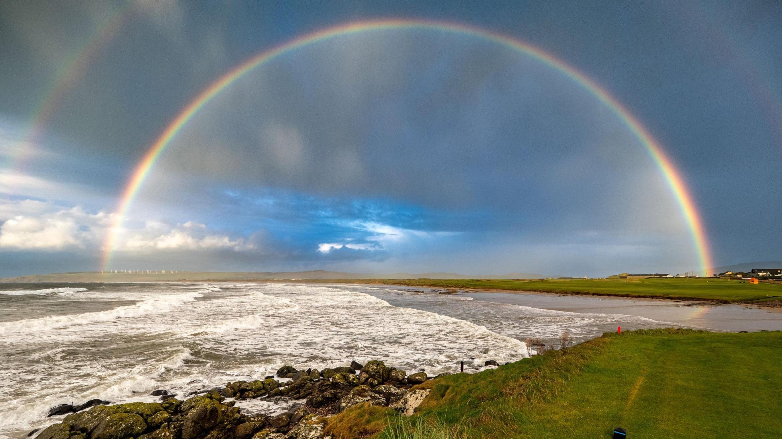 A sweeping rainbow arcs across a dramatic coastal scene, with waves crashing against dark, moss-covered rocks. The foreground features lush green grass leading to the edge of the shore, contrasting with the stormy sky above. Hints of blue sky peek through the clouds.