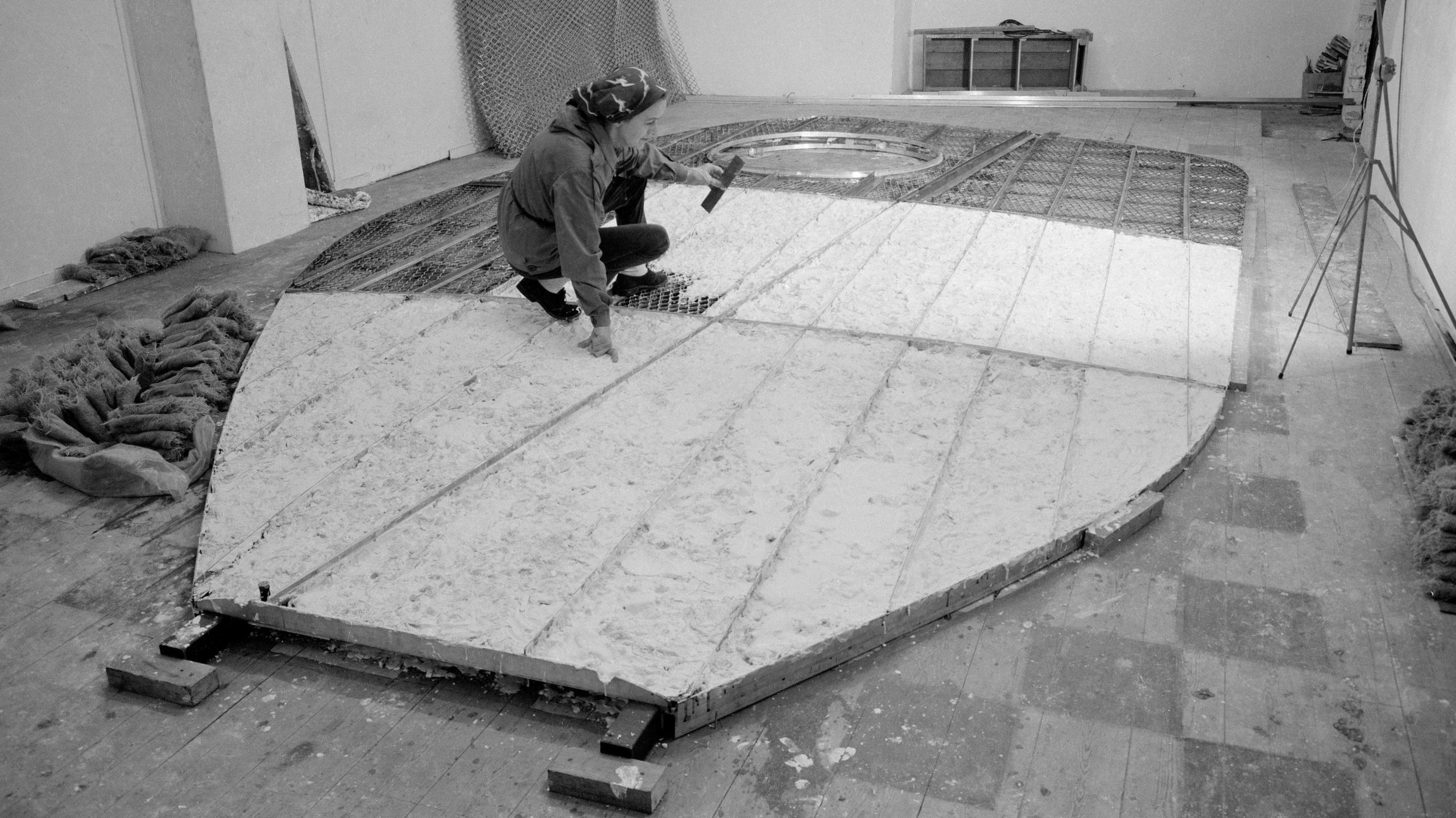 A black and white photograph of Barbara Hepworth working in the Palais de Danse on the prototype for Single Form for the United Nations in January 1961. The prototype is a large flat shape on the floor which she is crouching over. 