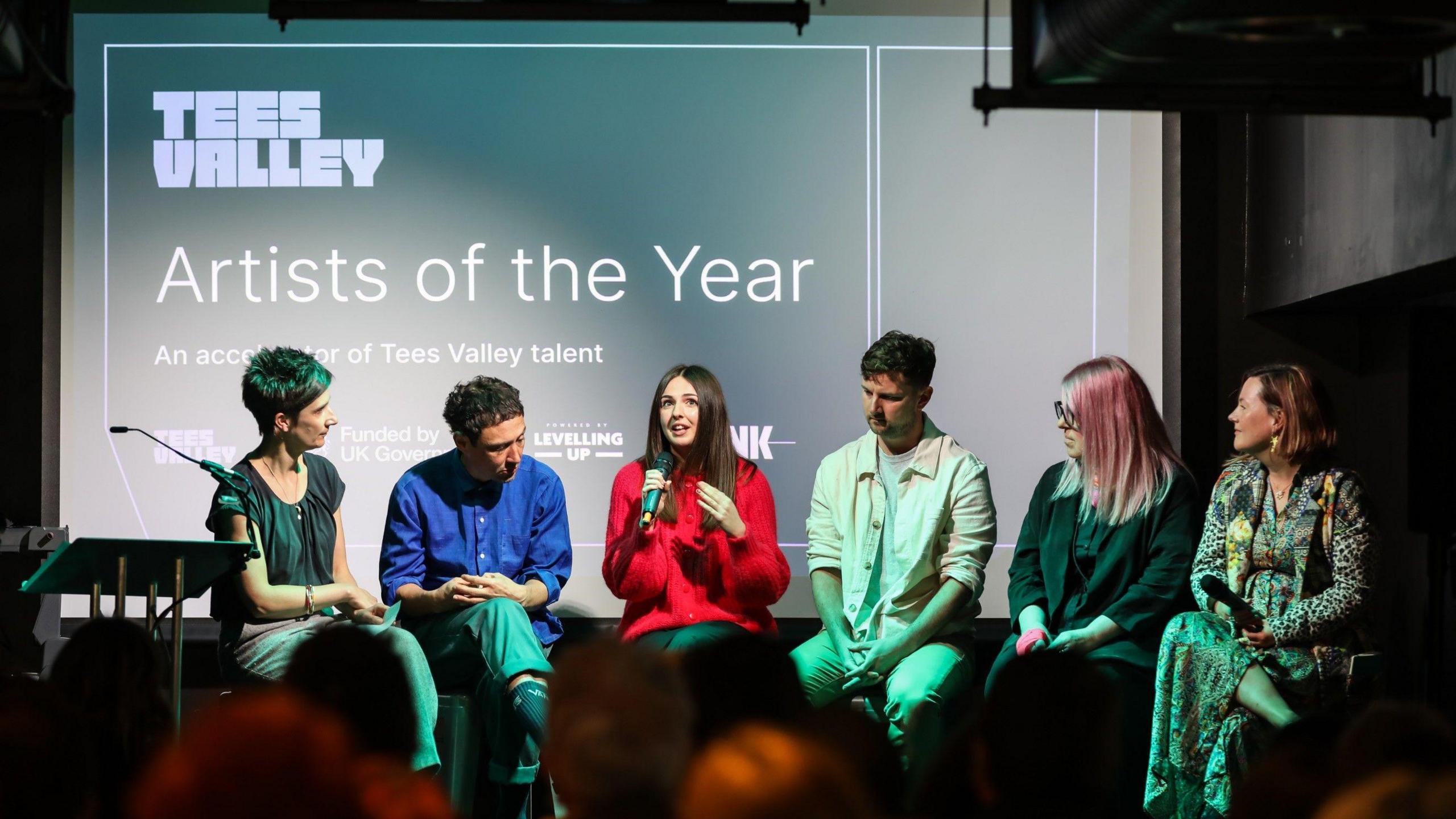 The five artists selected for the Artists of the Year programme in 2024 on stage during a talk about the scheme. They are joined by a host. A screen behind them displays the name of the programme and Tees Valley in a large font.