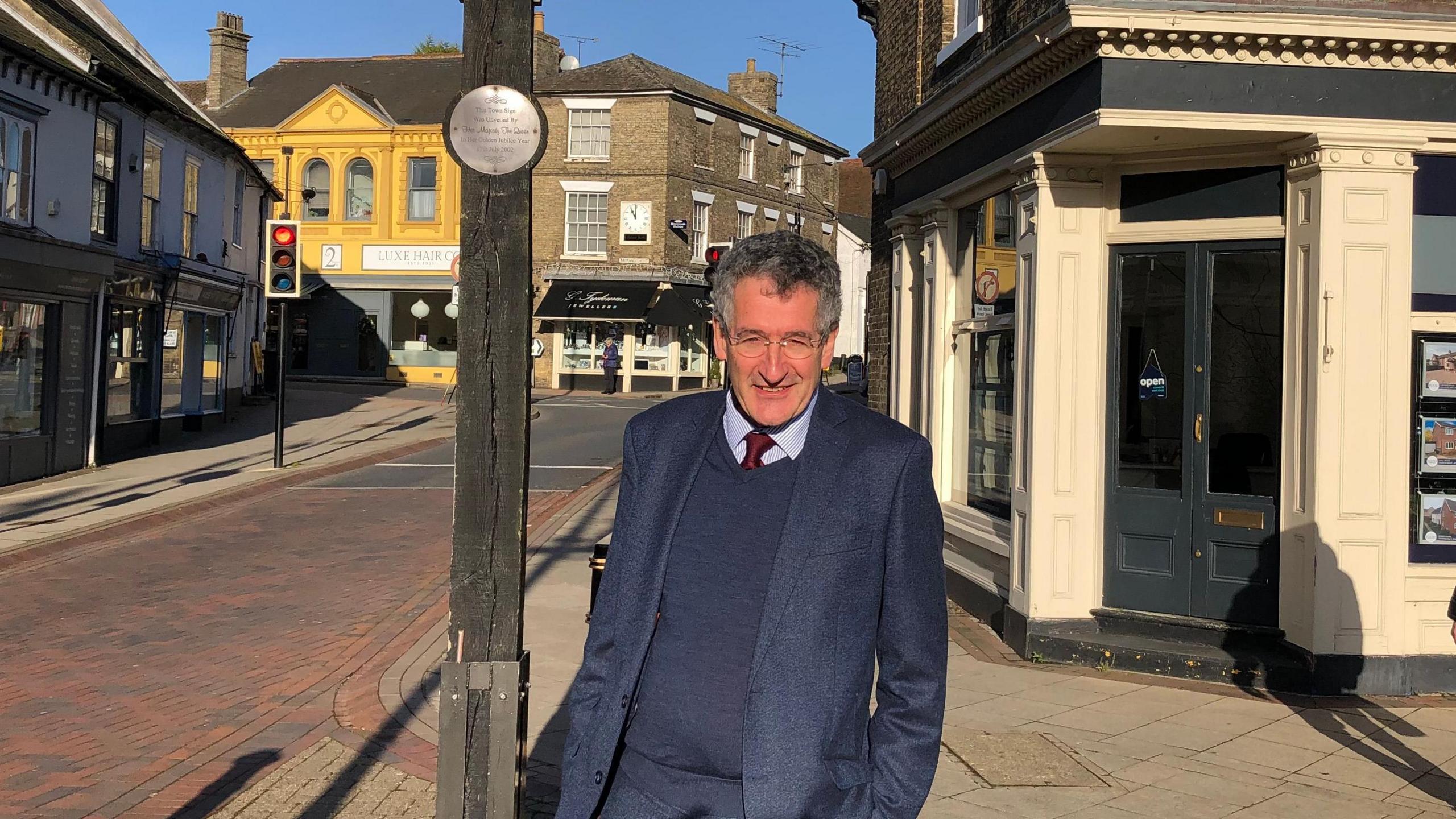 Suffolk MP Peter Prinsley wearing a tie and blue suit standing in the street. He has short dark curly hair and is next to a wooden post with a metal plaque on it. 