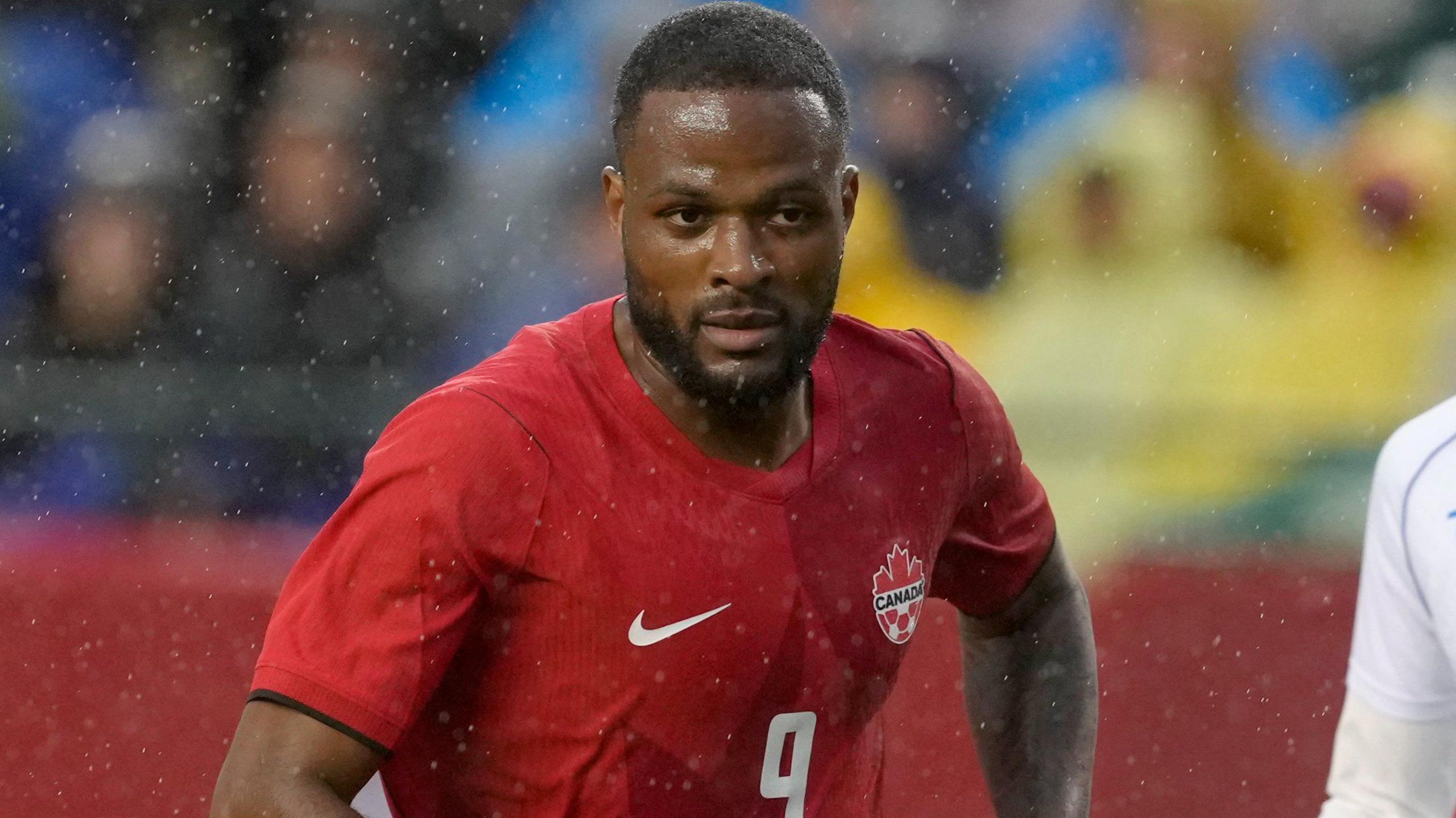 Cyle Larin looks straight-faced while in a game, wearing Canada's all-red home shirt with a white Nike tick and Canada logo.