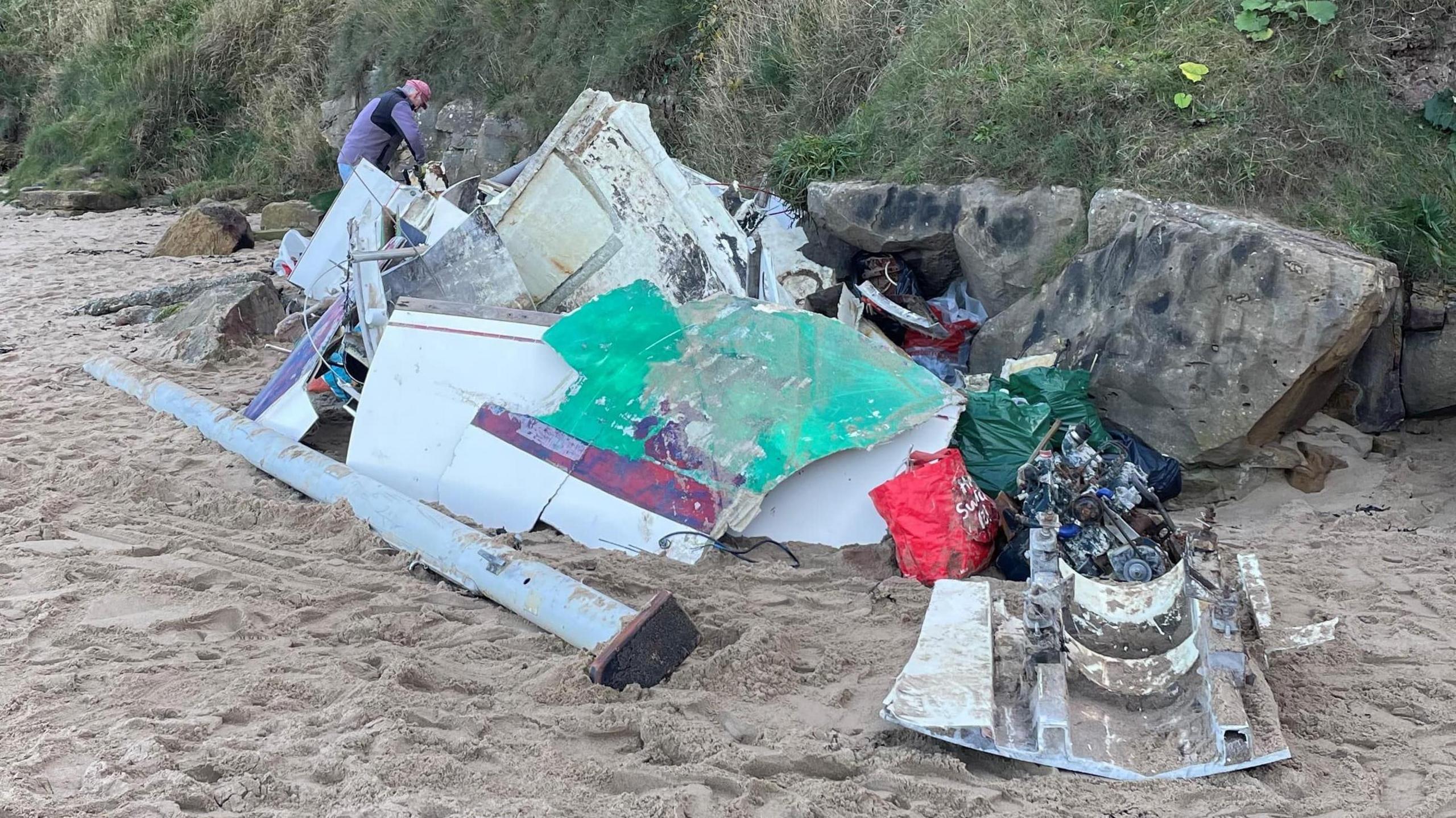 Man in purple top in the background of the photo with a pink cap cutting masses of debris on a beach. The debris is made up of green and white pieces of a boat covered in sand. 