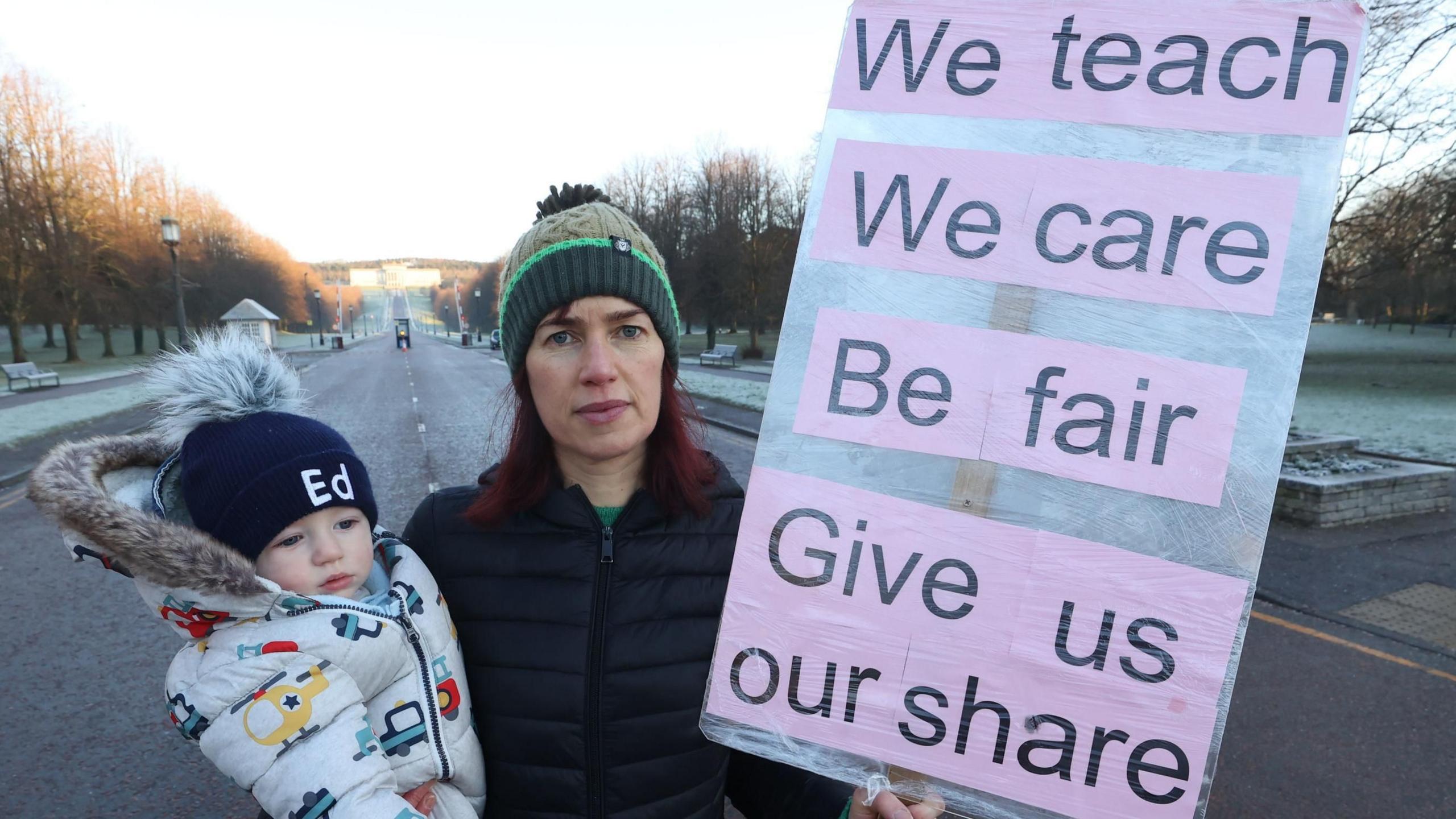 Woman wearing a green bobble hat and black padded coat. She is holding a baby in one arm wearing a white padded parka with places and cars on it, they also have a navy bobble hat on with Ed on the front. In her other hand, she is holding a sign reading 'We teach, We care, Be fair, Give us our share'. Stormont can be seen in the background.  