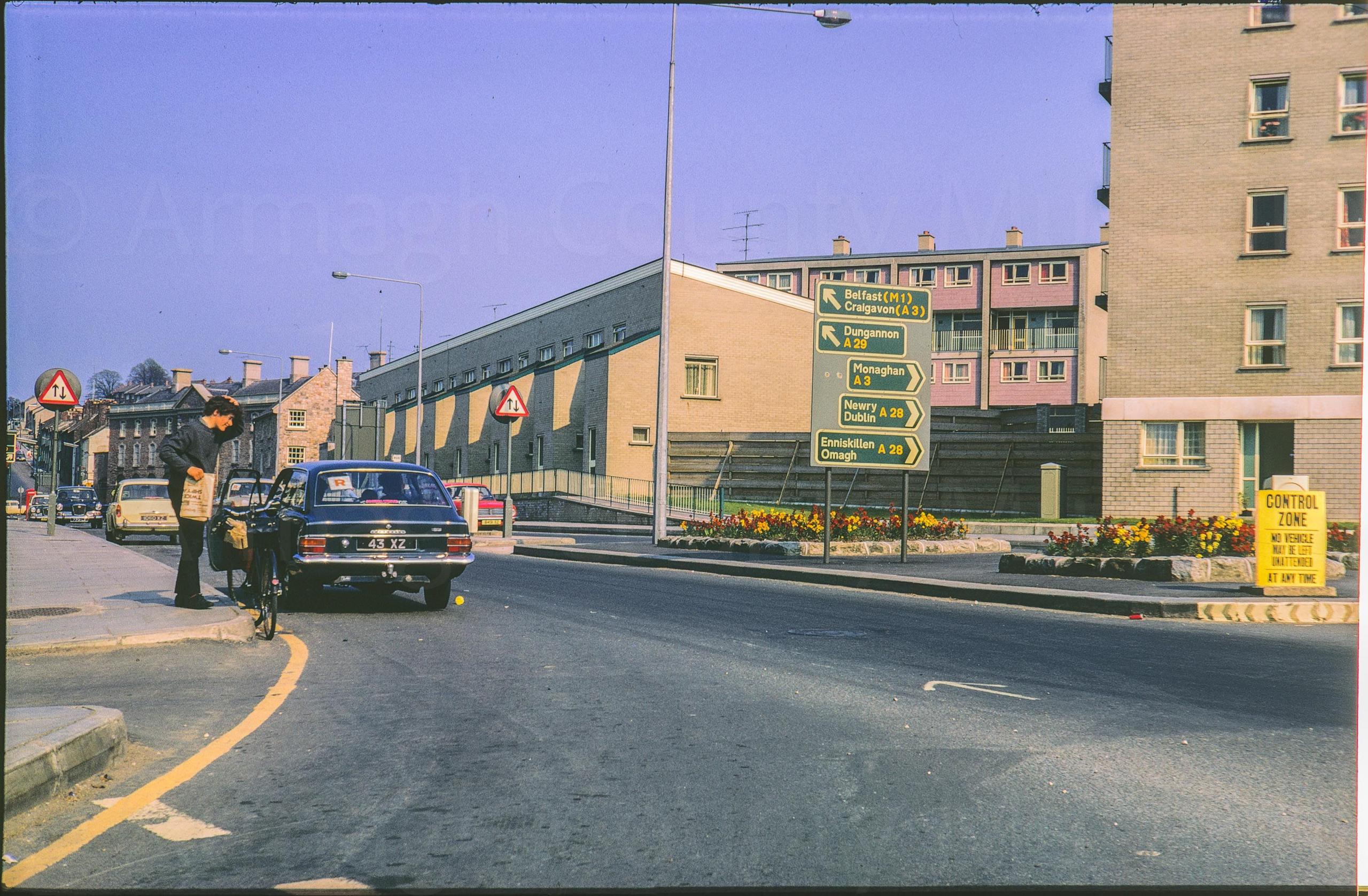 A digitalised photo from a film of a crossroads street with a block of flats.  A young man stands in the foreground beside a bicycle holding a newspaper with the prison visible in distance behind him.