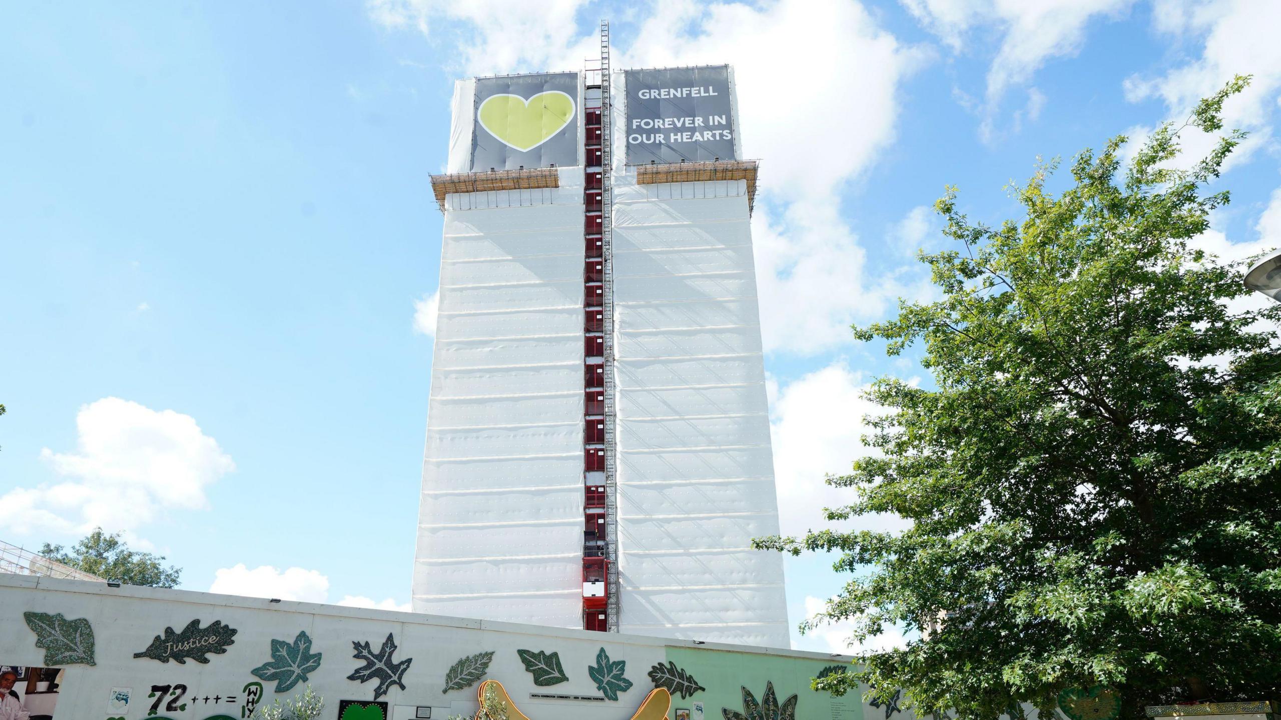 Image shows Grenfell Tower and the a wall full of memorials and messages on a sunny day, with blue sky and clouds