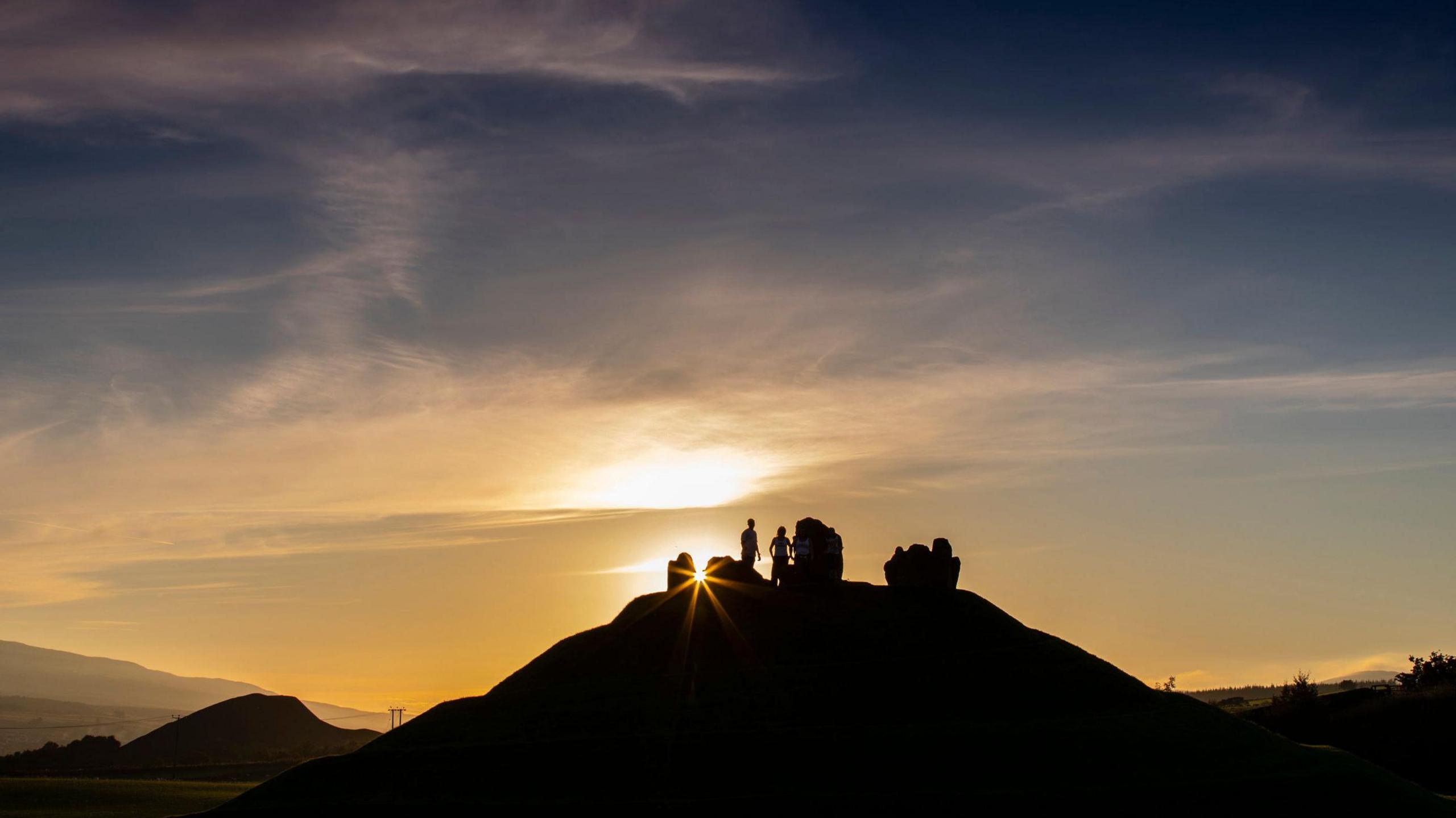 A sunrise at the Crawick Multiverse with light breaking through the rock artwork on top of a hill