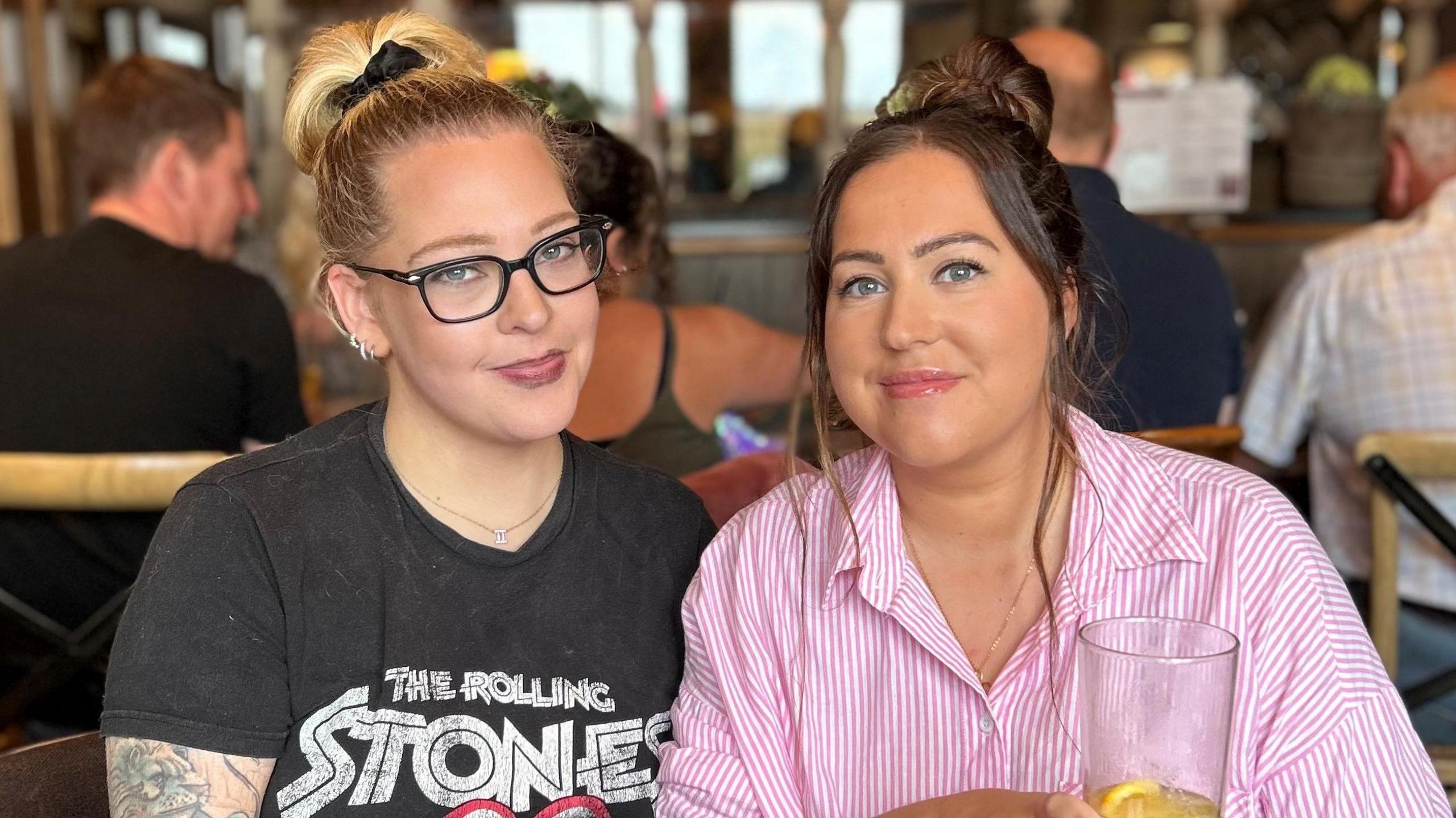 Becky and Nikki Roberts, sitting down in a bar and smiling at the camera. Becky has blonde hair in a pony tail, black glasses, three loop earrings in her right ear and a tattoo on her arm. She is wearing a black Rolling Stones T-shirt. Nikki has brunette hair in a bun and is wearing a pink and white striped shirt. She is holding a drink.