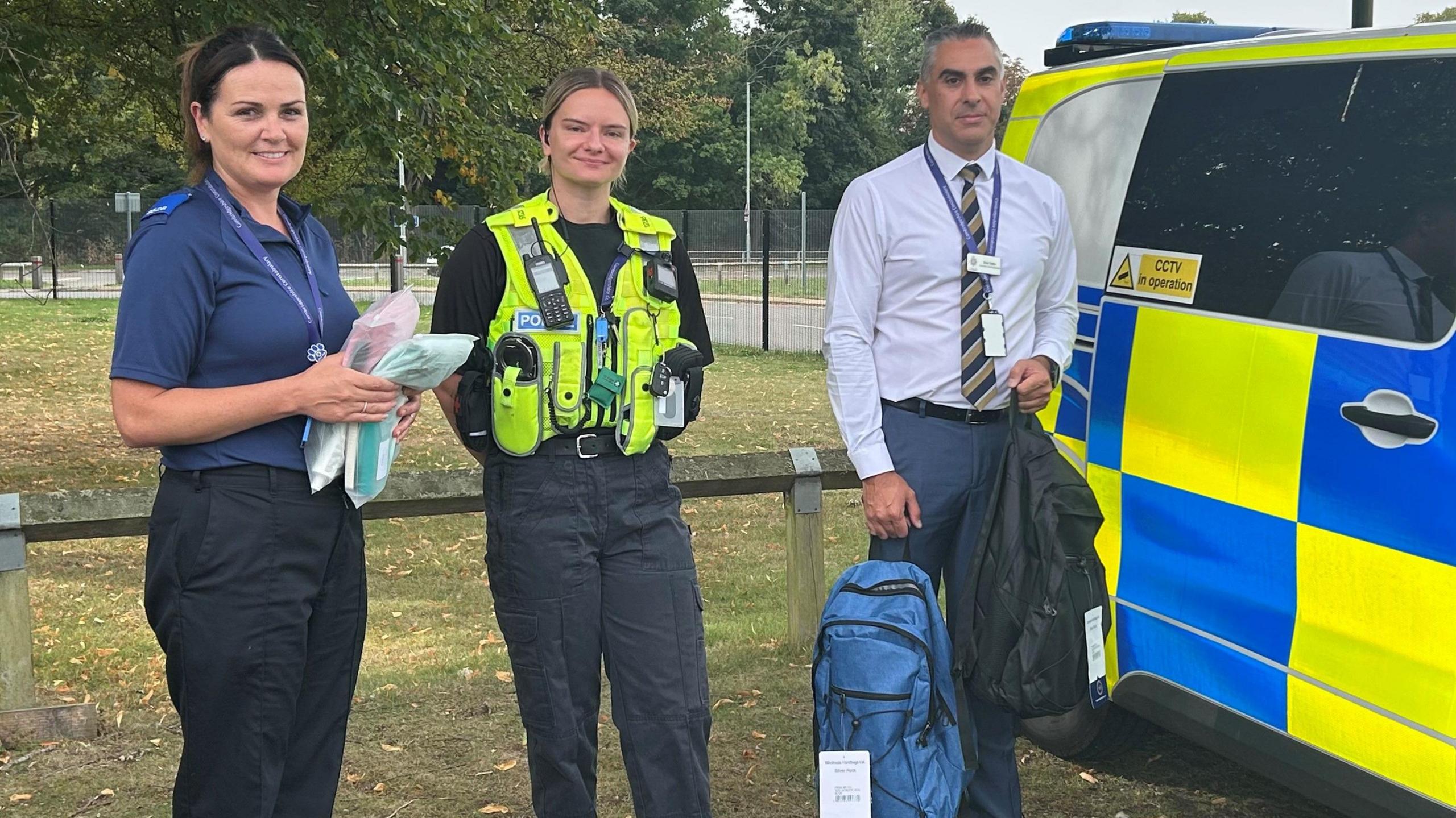 Two women officers - one wearing a blue T-shirt and black trousers and another in a green police jacket - and Det Ch Insp Taylor wearing a white formal shirt , a striped black and gold tie and grey trousers- holding a blue grab bag- standing next to a police car.