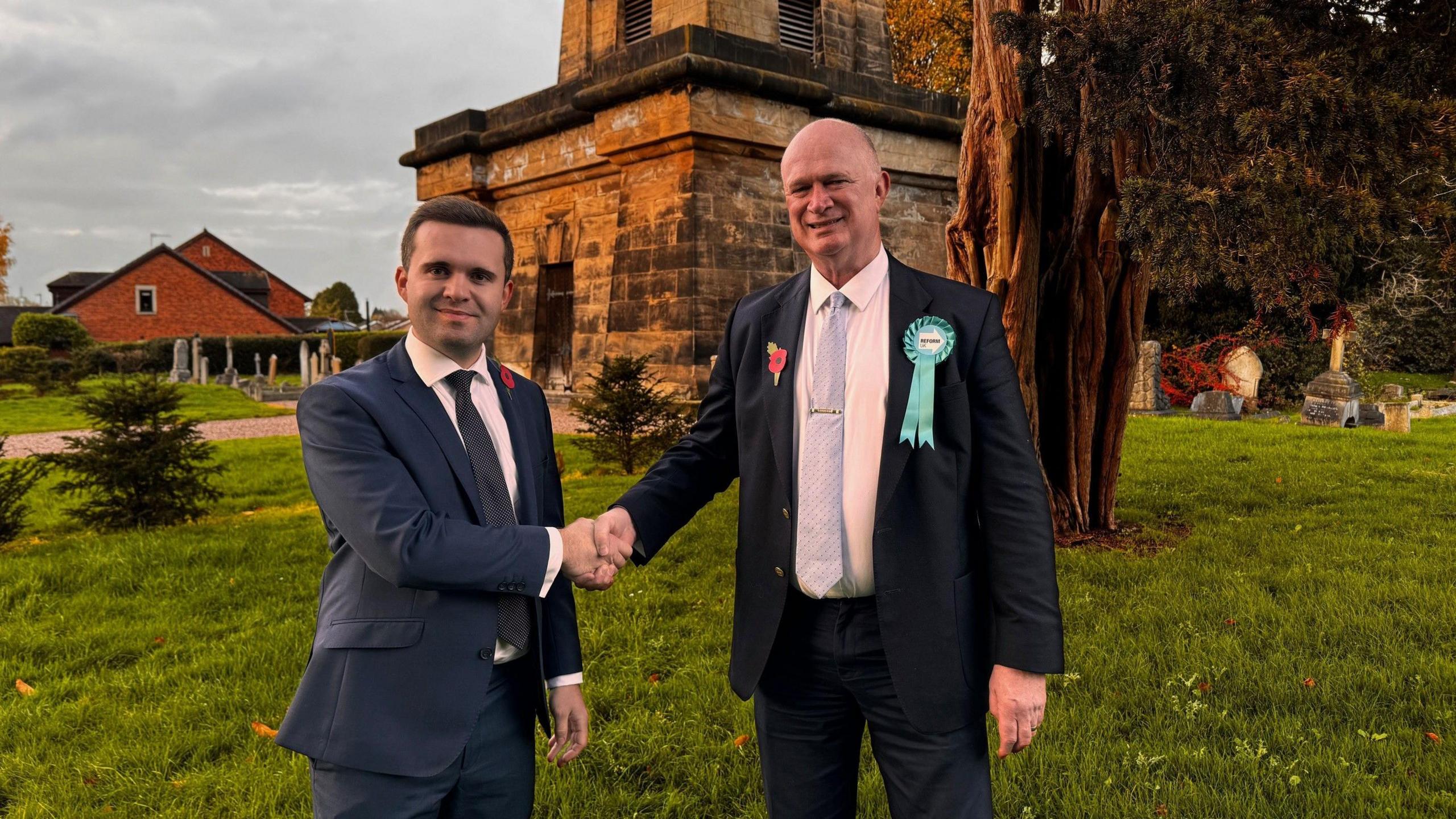 Two men wearing dark suits are seen shaking hands. They are stood in what appears to be a churchyard.