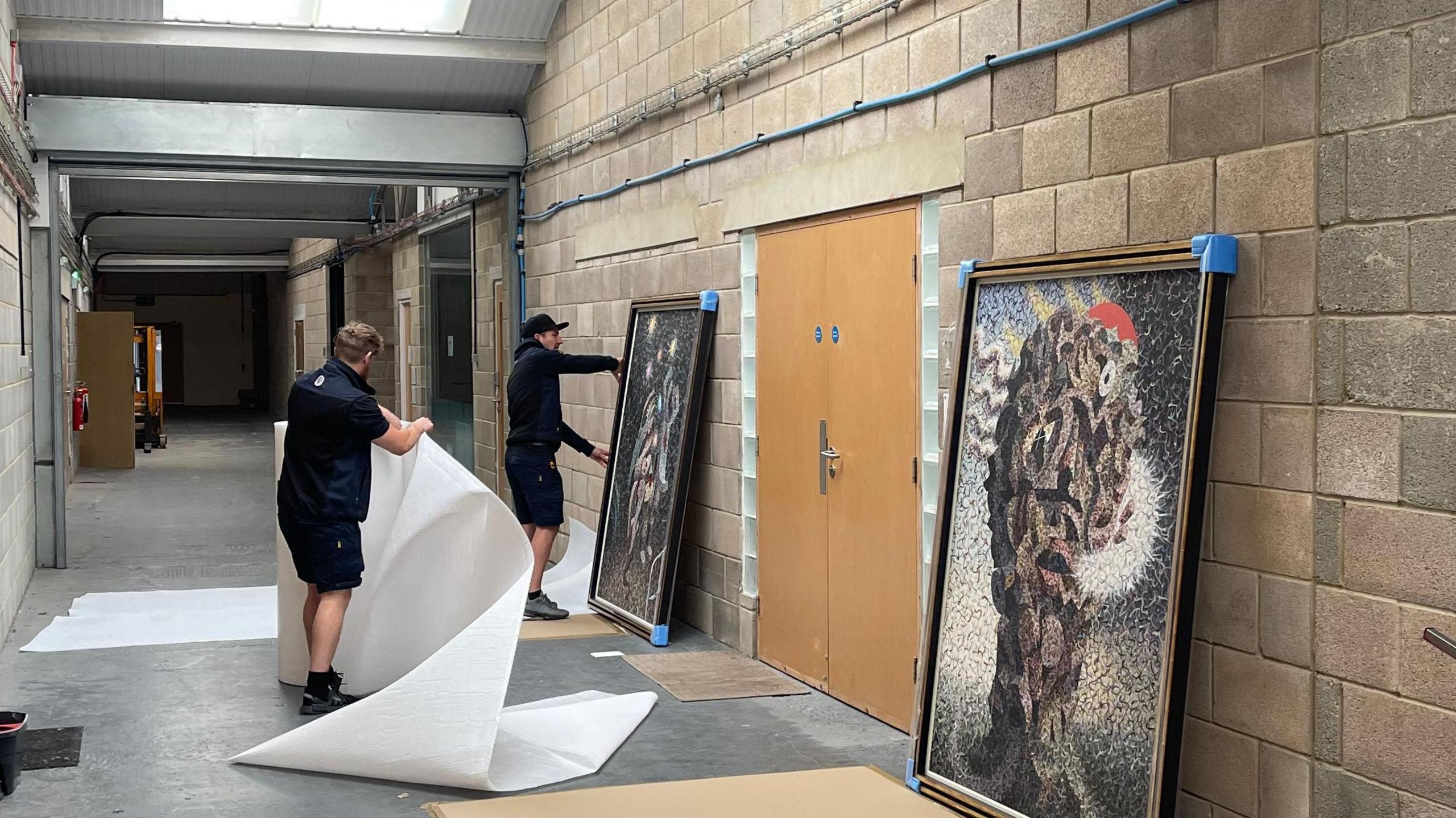 Two people in a warehouse-style corridor with two large paintings leaned against a wall