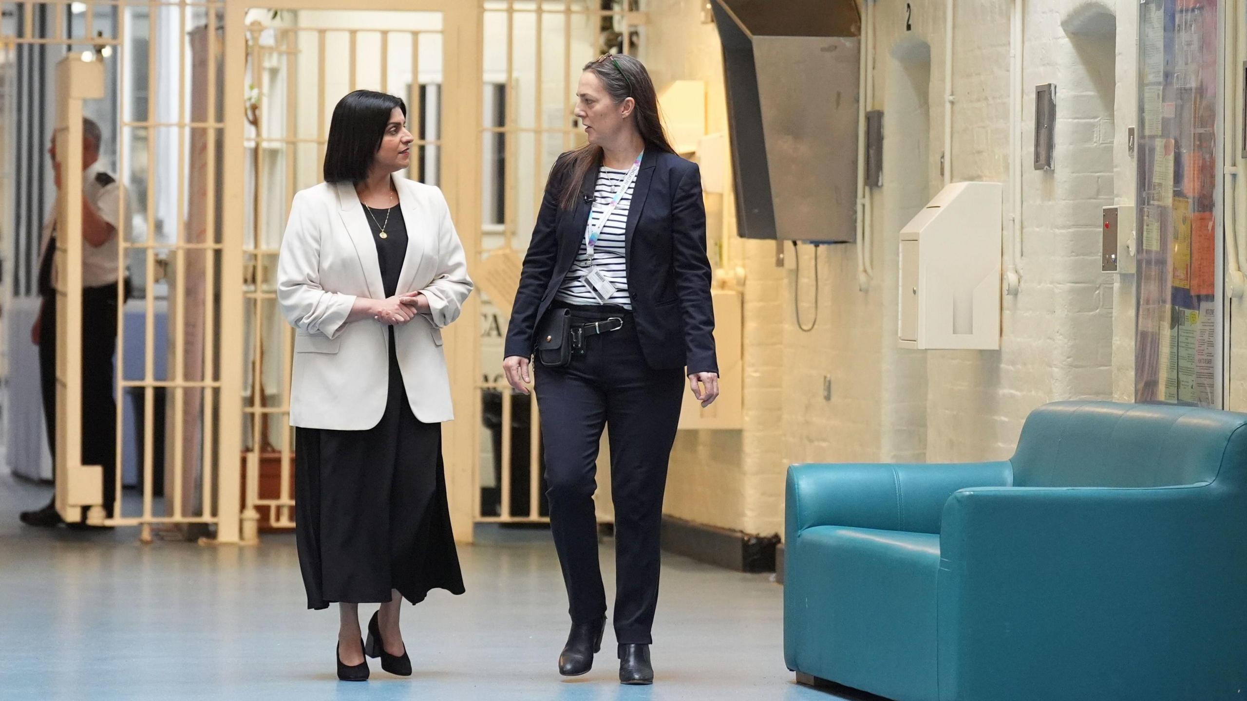 Shabana Mahmood visits HMP Bedford, alongside the prison's governor, Sarah Bott. The pair walk along a while corridor and in the background there is a prison officer holding open a white gate.