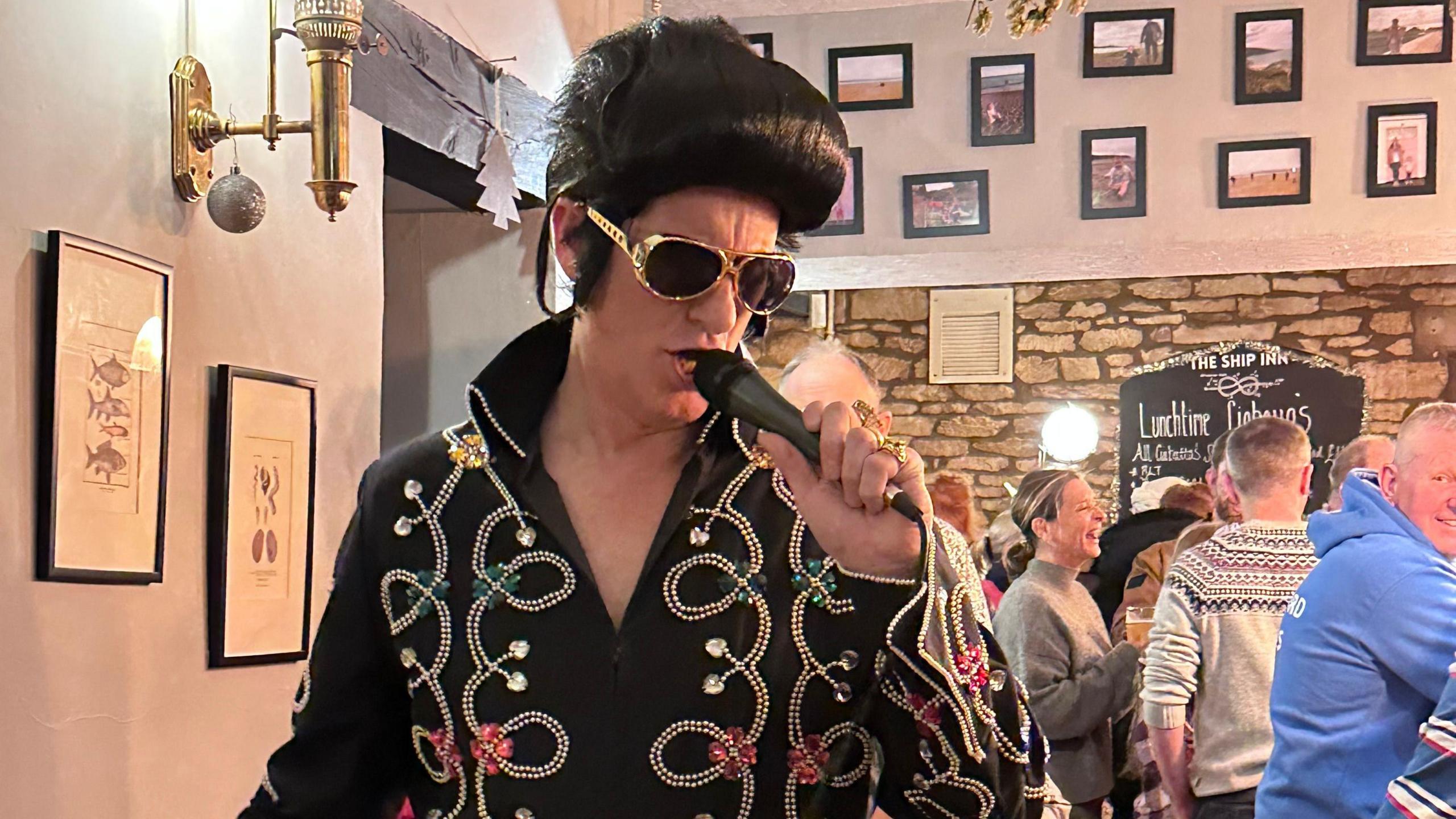 A man dressed as Elvis sings into a microphone in a pub as patrons stand around, drinking and laughing