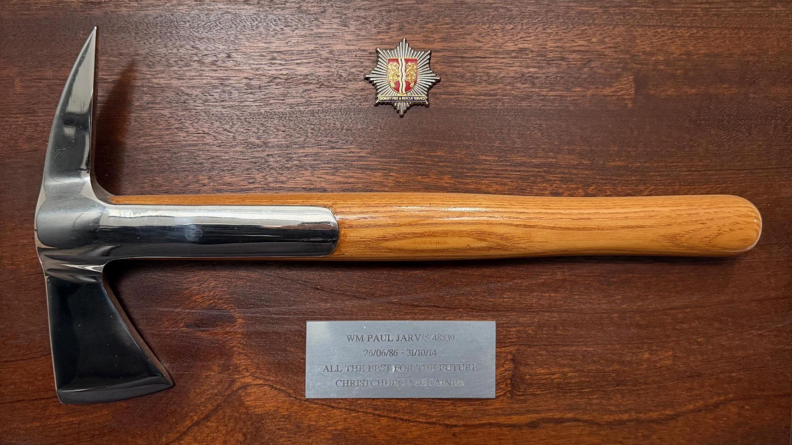 A ceremonial fire axe mounted on a wooden board with a metal plaque commemorating nearly 30 years of service as a retained firefighter by Paul Jarvis