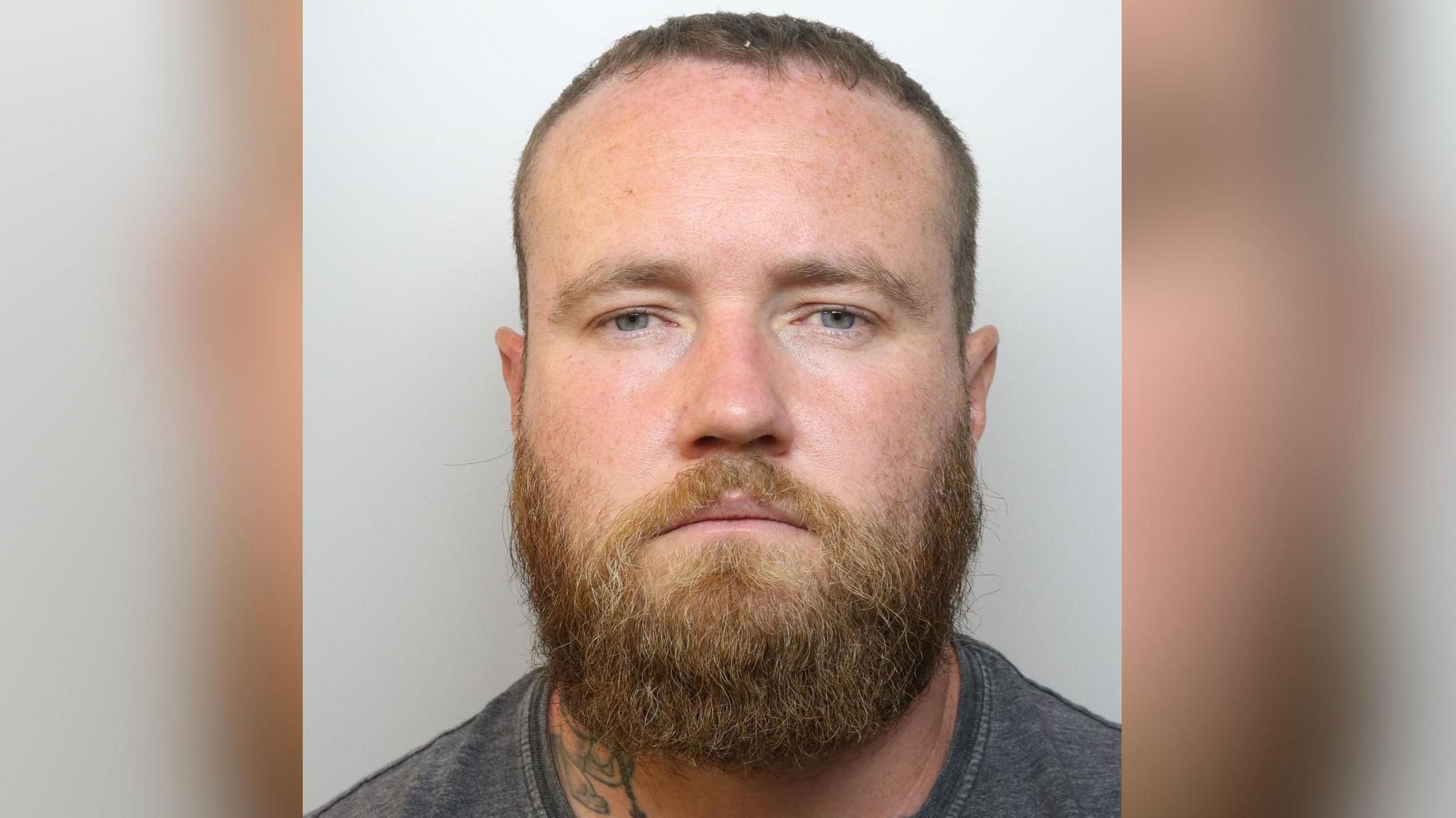 A police mugshot of Colin Brown, who has blue eyes, a ginger beard and moustache and short brown hair. He is wearing a grey T-shirt.