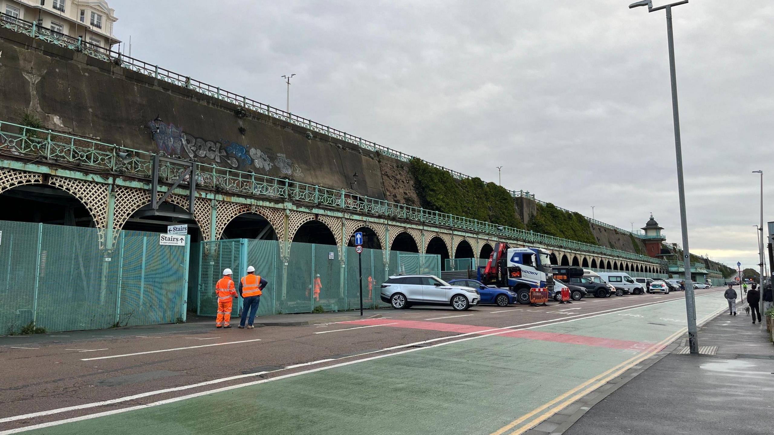 A picture of the fenced off Madeira Terrace taken from ground level, showing four workers in hi vis clothing and a row of cars parked in front of the structure.