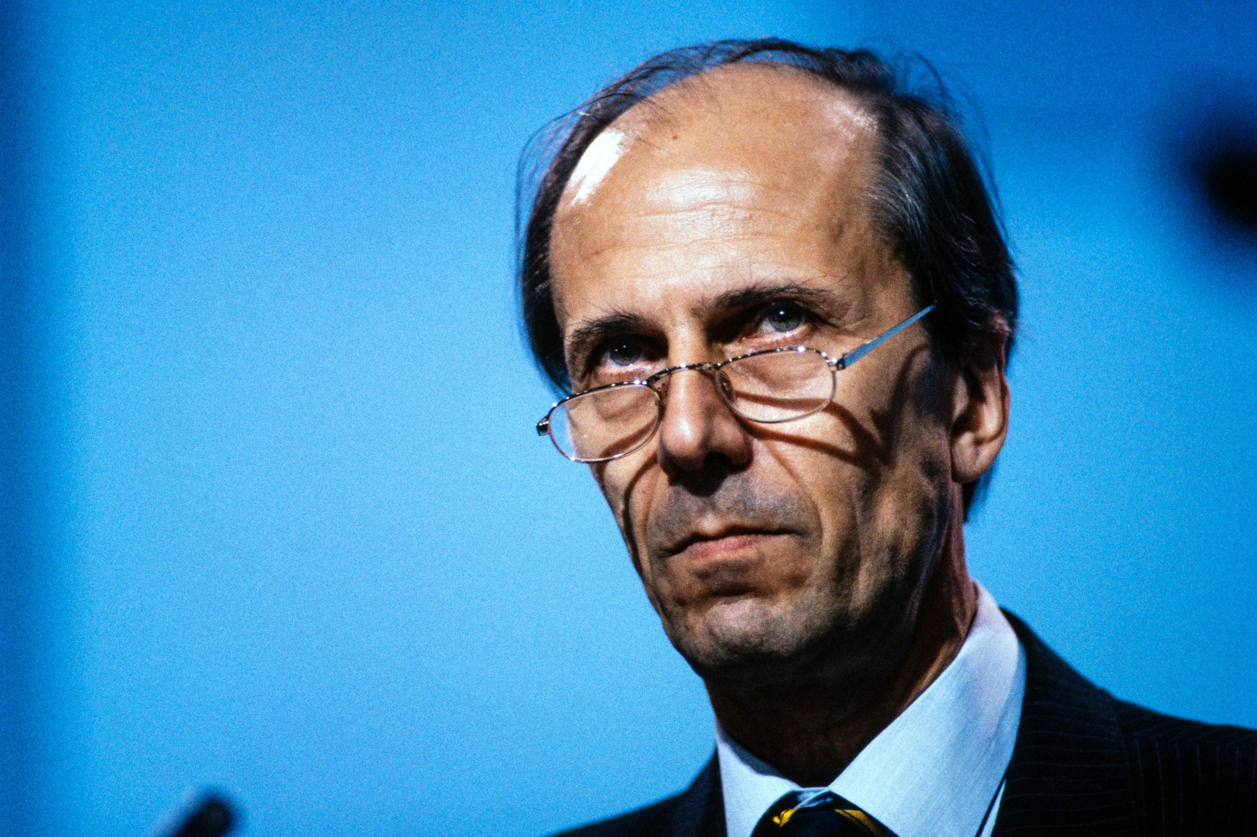 Norman Tebbit ka veshur një kostum me vija të errëta, një këmishë të bardhë dhe një kravatë me motive me vija diagonale në të verdhë dhe të zezë. Sfondi është blu i plotë, dhe një pjesë e një mikrofoni është e dukshme në anën e majtë të imazhit, duke sugjeruar një mjedis formal për fjalime ose prezantim.
