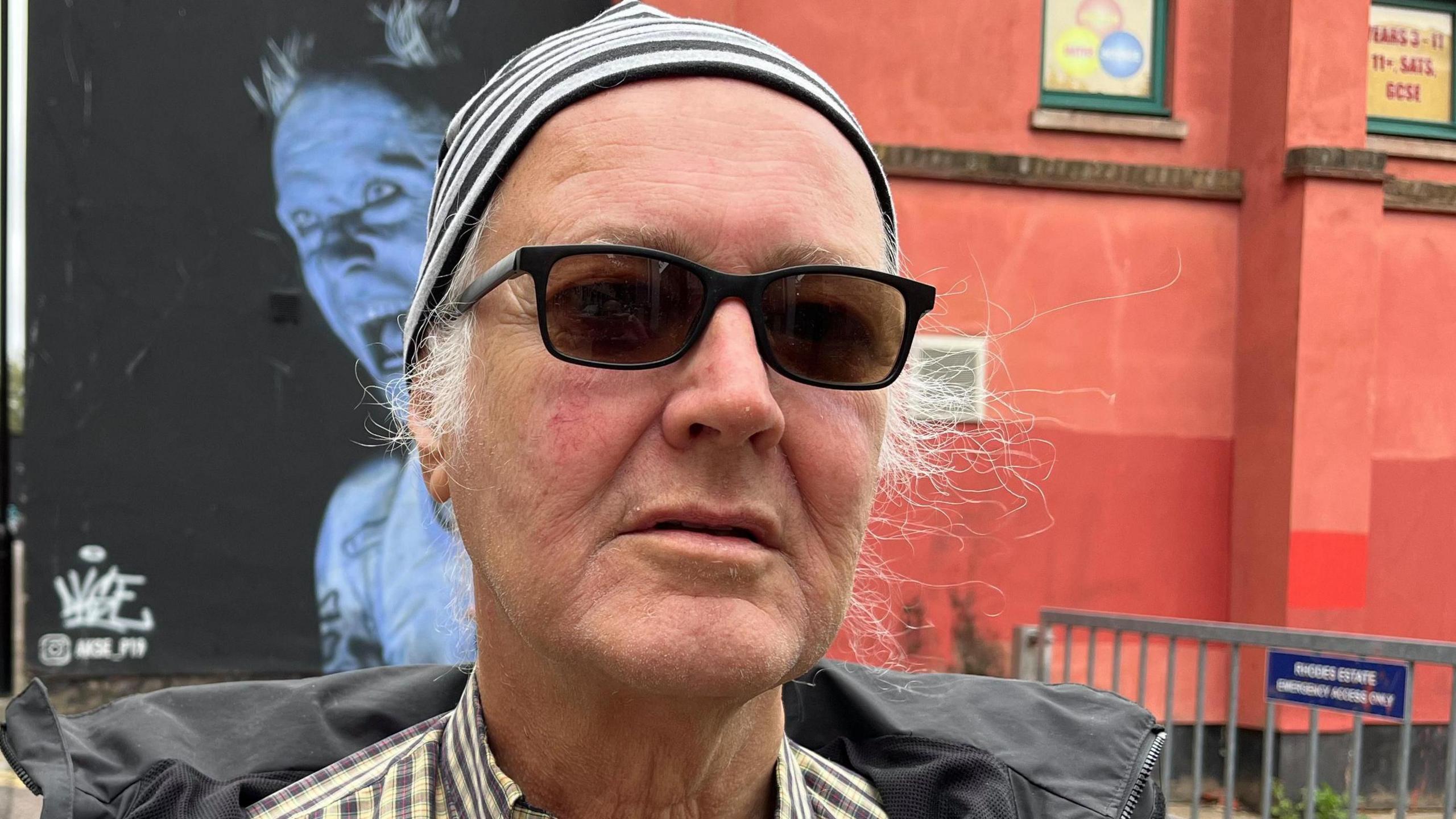 A man looks at the camera. He is wearing a hat and sunglasses.