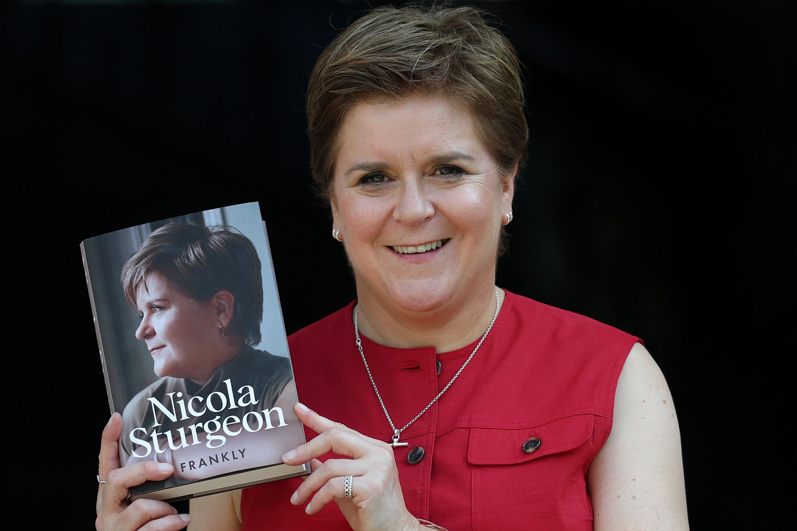 Nicola Sturgeon, who  has short brown hair combed in a side shed and is wearing a sleeveless red blouse, holding a copy of her memoir, Frankly.