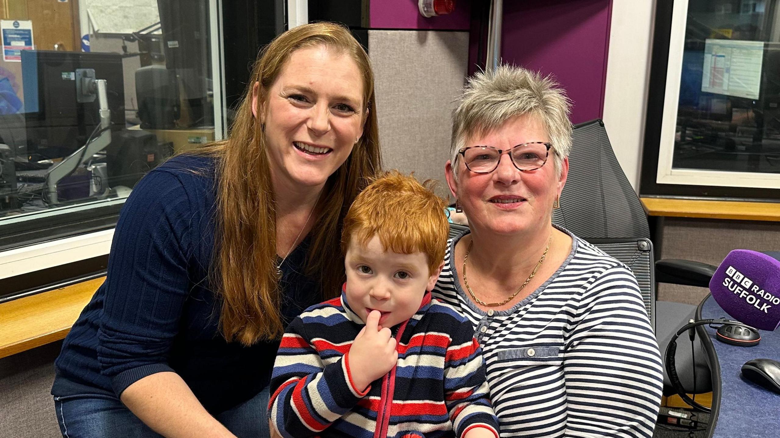A head and shoulders image of three people pictured in a radio studio. A woman with grey short hair and glasses, and wearing a dark grey and white striped top, is seated, with a three-year-old boy on her lap. He has red hair and is wearing a blue, red, black and and white striped fleece. The tip of his right index finger is in his mouth. Crouching to his level, to his right, is a woman with long red hair, dressed all in blue. All three are looking at the camera.