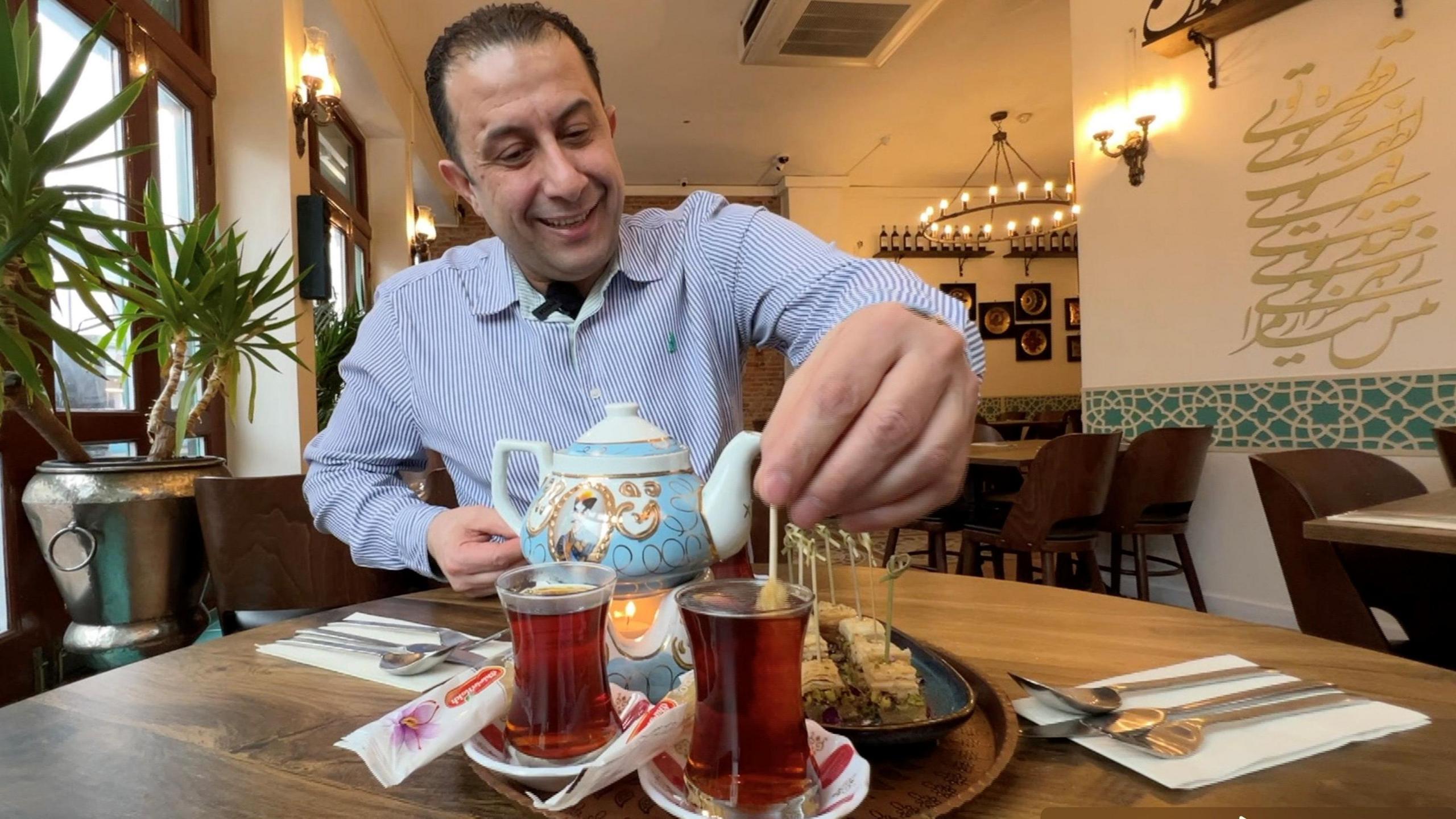 Massood Khamloo serves traditional Iranian tea in his restaurant, Shiraz, on Cowley Road, Oxford. Smiling, he stirs a black tea in a glass with a saffron sugar stick. A plate of baklava is next to the tea. 