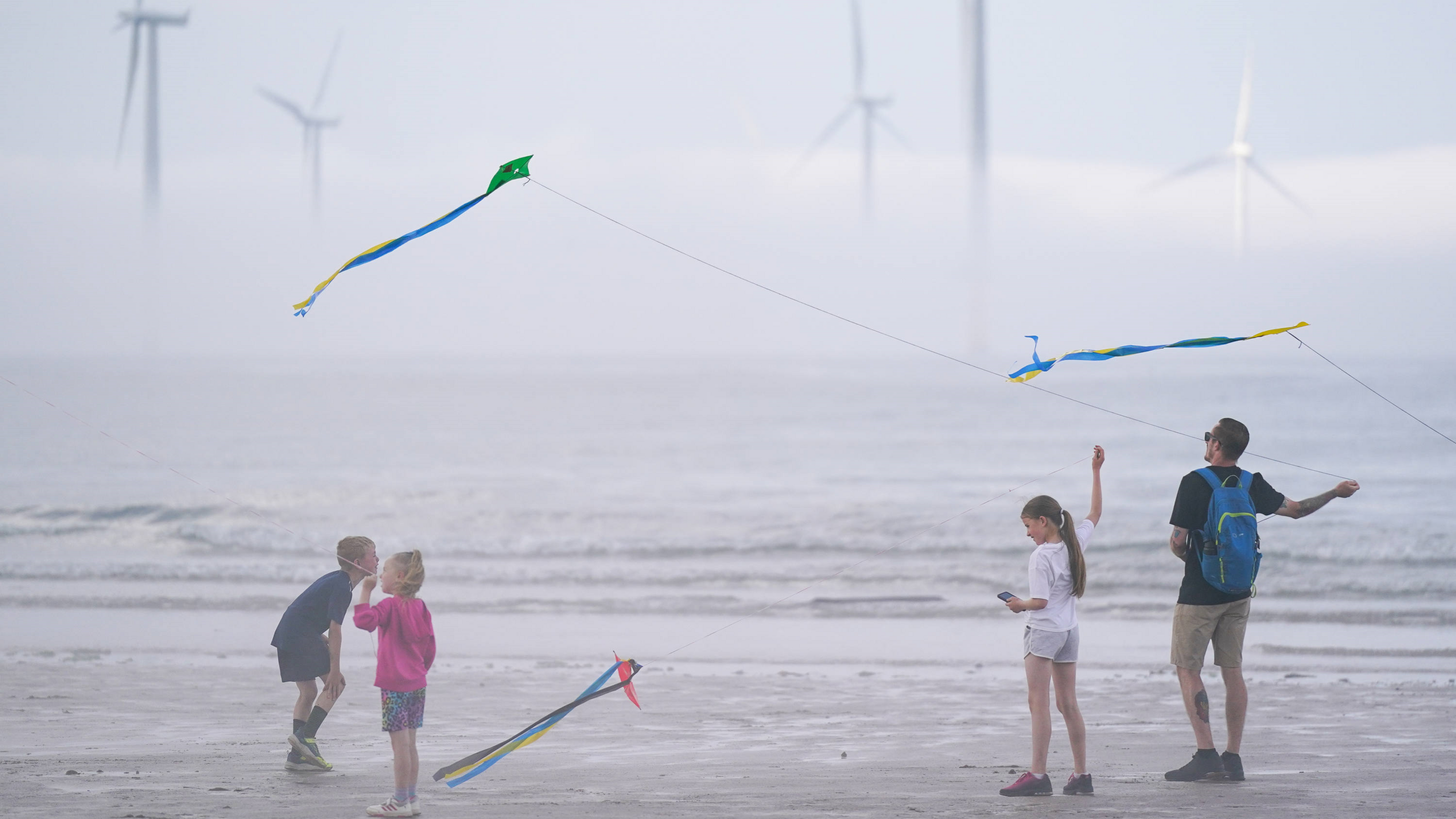 A family flying kites have been pictured standing on the shoreline with offshore wind turbines emerging from the fog in the background.