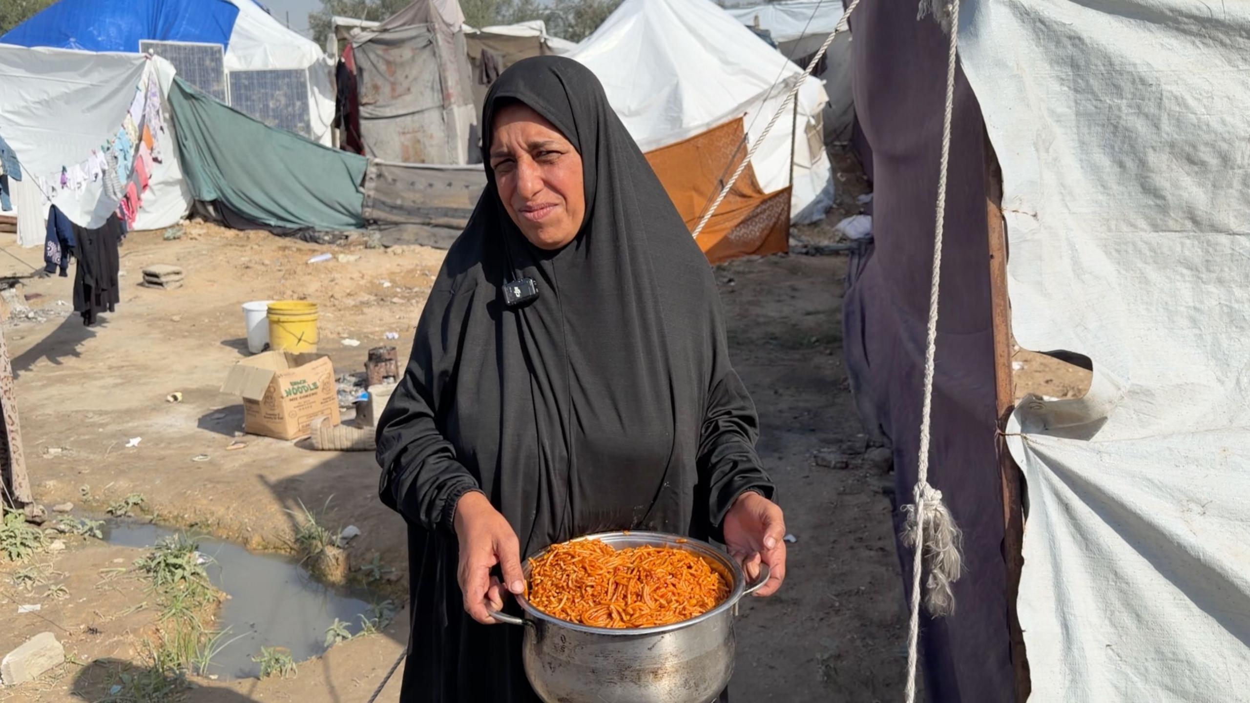Aida Salha holds a pan full of pasta from an Anera community kitchen in central Gaza. Tents can be seen around her and she is wearing a black dress and headscarf