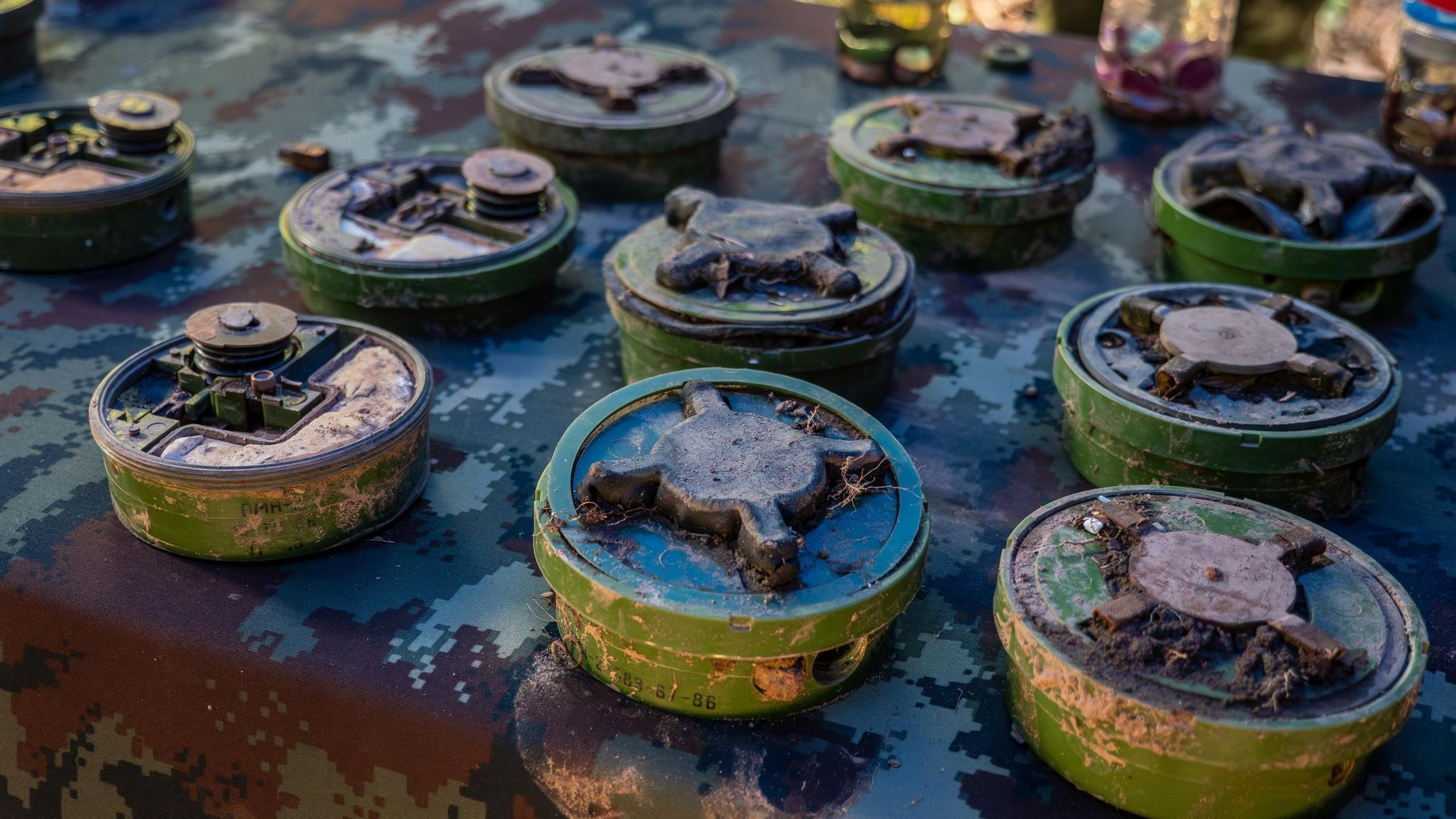 Ten landmines are laid out on a table covered with a camoflauge print cloth. The mines are green and have a cross pattern atop them. Some have soil on them, indicating that they have been recently dug up.