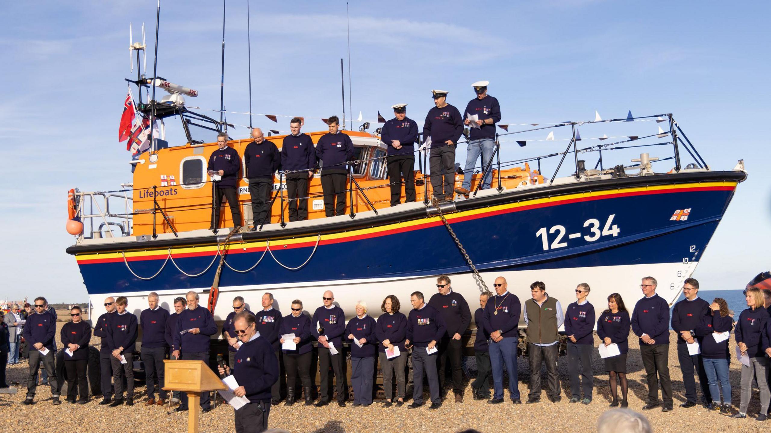 The Freddie Cooper all-weather lifeboat - an orange and navy blue vessel - on a beach. Dignitaries and RNLI staff can be seen standing on and in front of it.