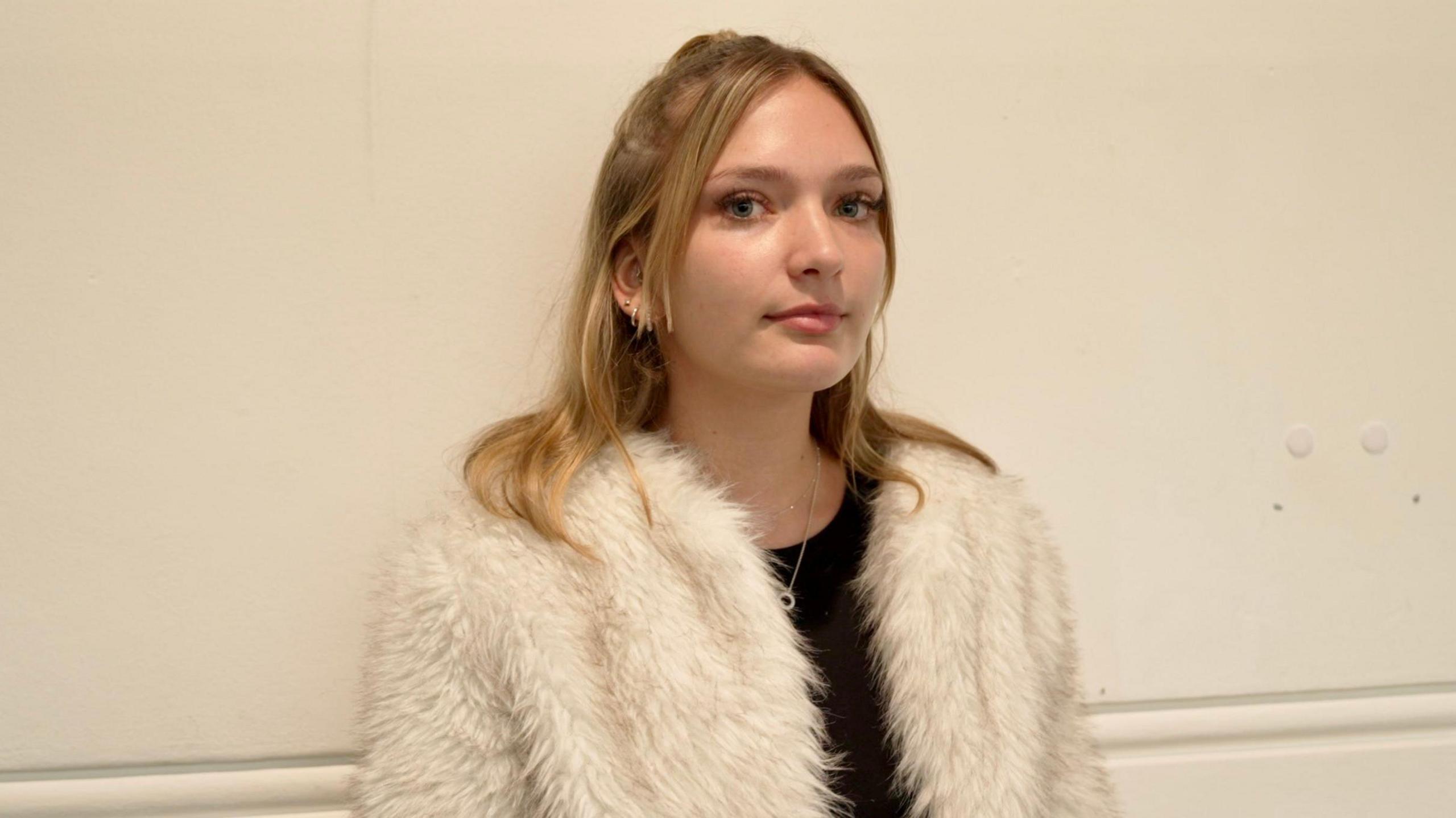 A young woman wearing a white fur coat over a black top. She is stood in front of a white wall.