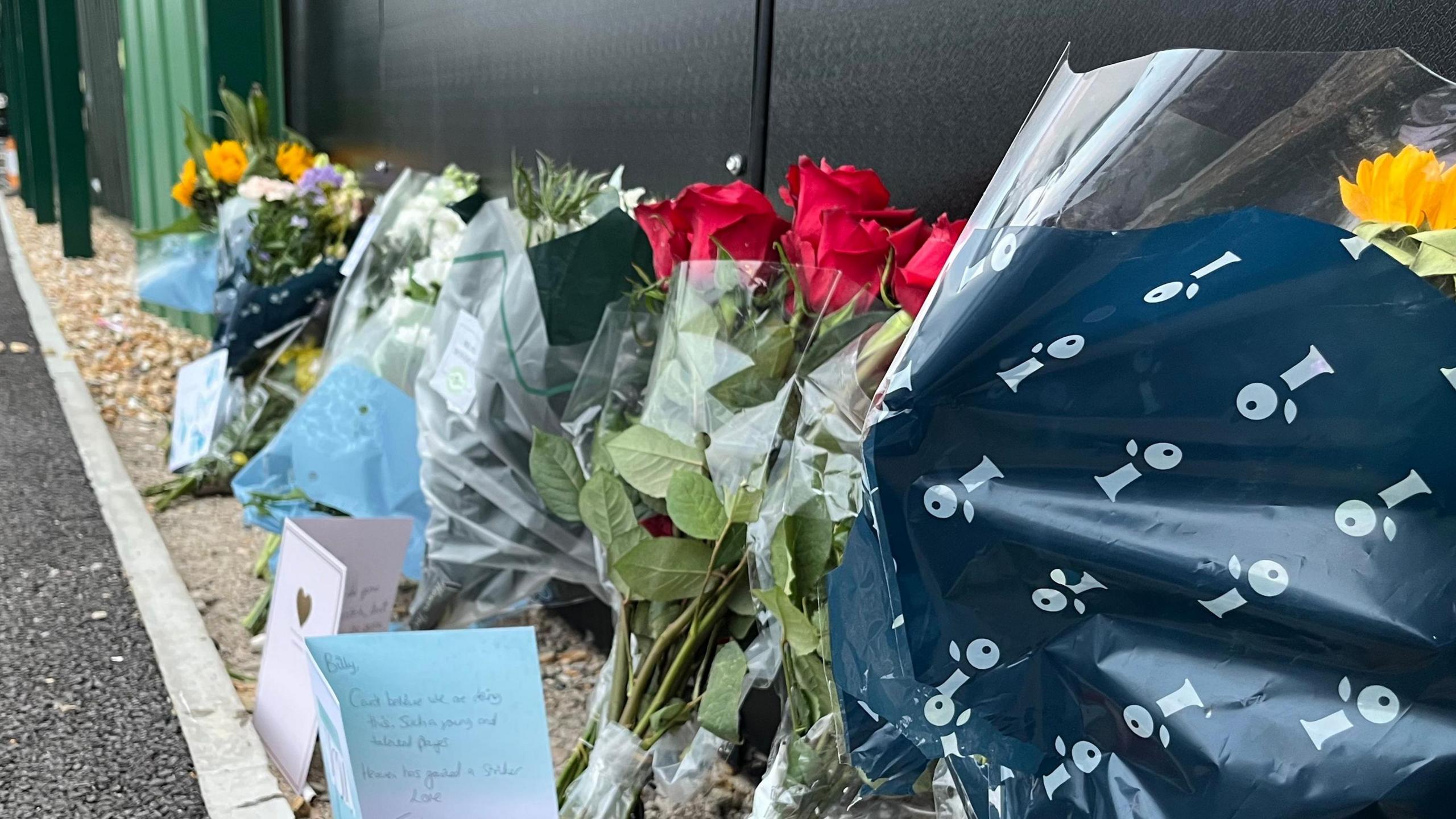 A number of floral and card tributes left for the late Billy Vigar at Chichester City football club. Six separate floral tributes can be seen in a row alongside three cards. 