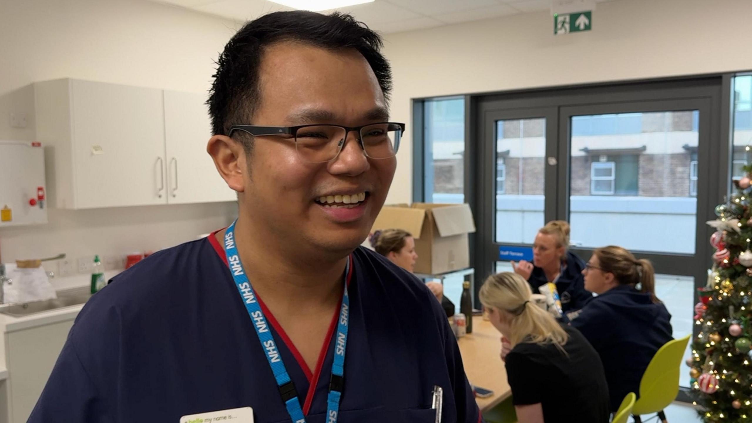 A man wearing glasses and a blue nurses uniform smiles as colleagues sit behind him in the staff rest area. In the background is a Christmas tree and patio doors onto a terrace. 