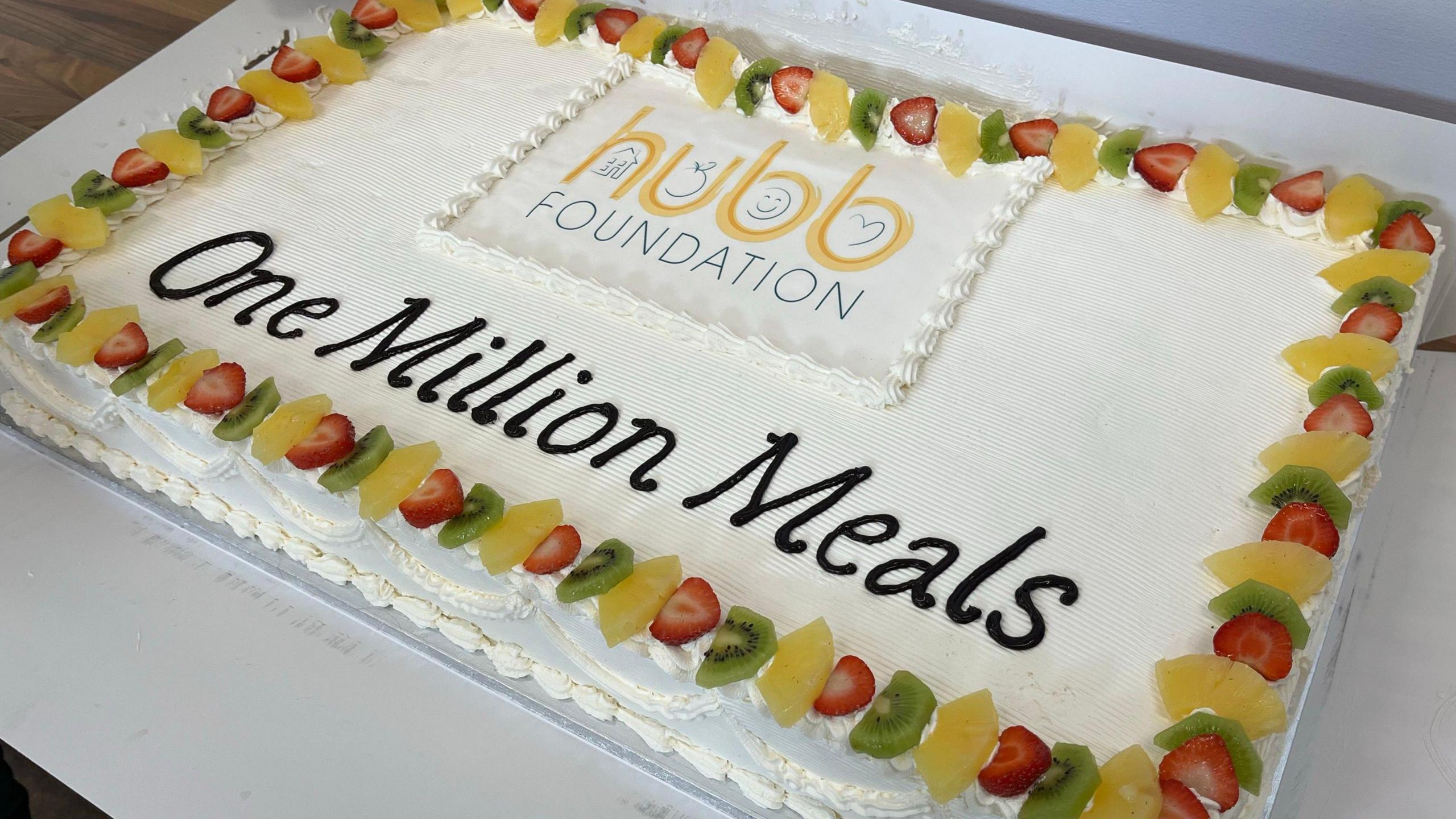 A white, rectangular cake with slices of strawberries, orange and kiwi fruit around the border with "Hubb Foundation , one million meals" written in the middle. 