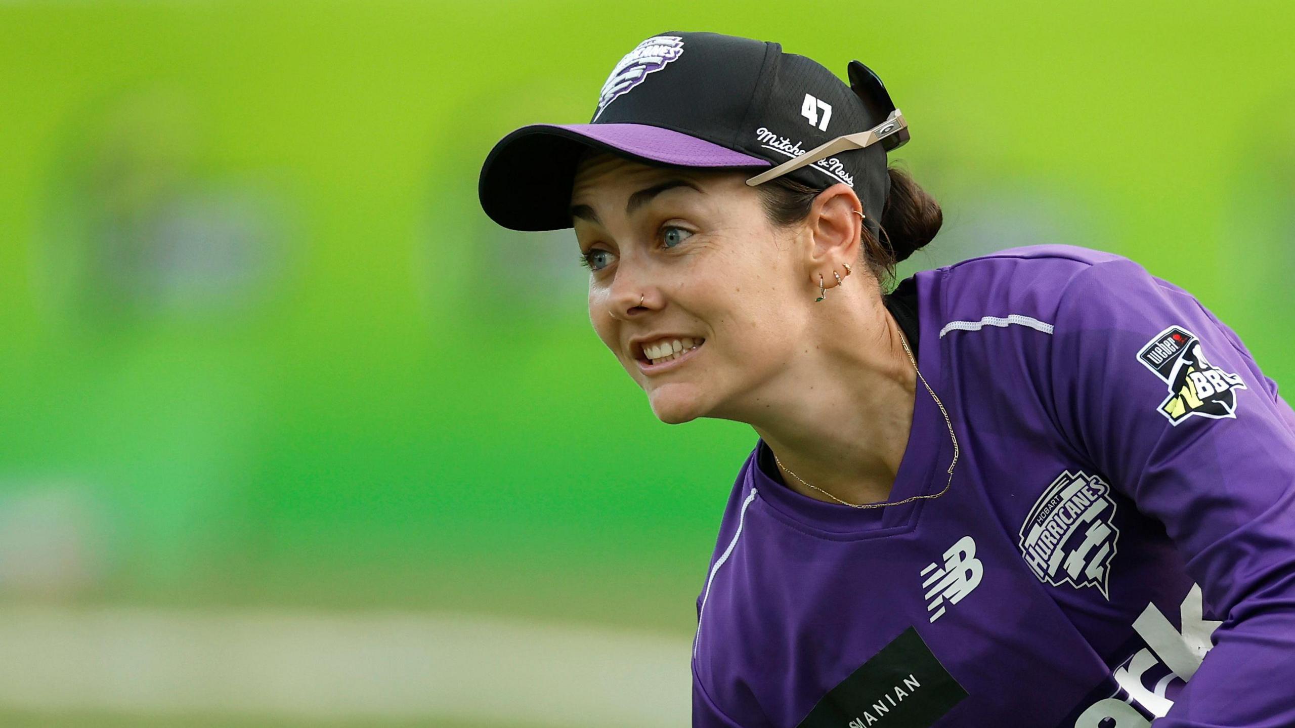 Heather Graham in action wearing a purple long sleeve kit and a black cap with a purple peak. Her sunglasses are on the back of her cap and she has a long gold chain around her neck.