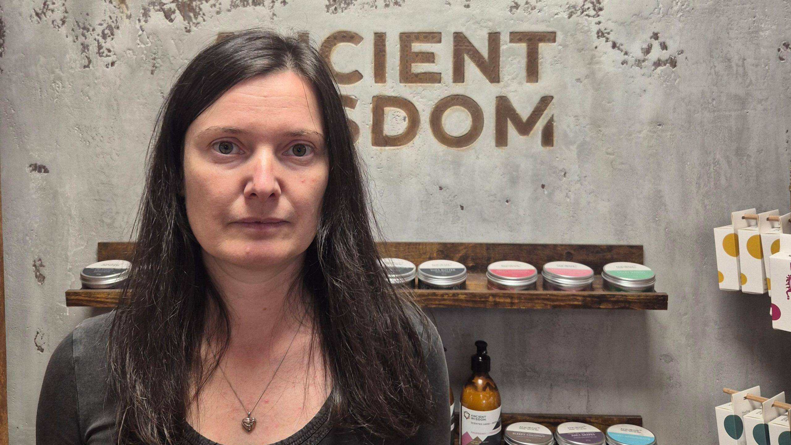 Katerina Buchy, a woman with long dark hair, wearing a dark grey top and a necklace, looks at the camera. The words "Ancient Wisdom" are written on the wall behind her in block capitals, and below that there is a shelf with cosmetic products on it.