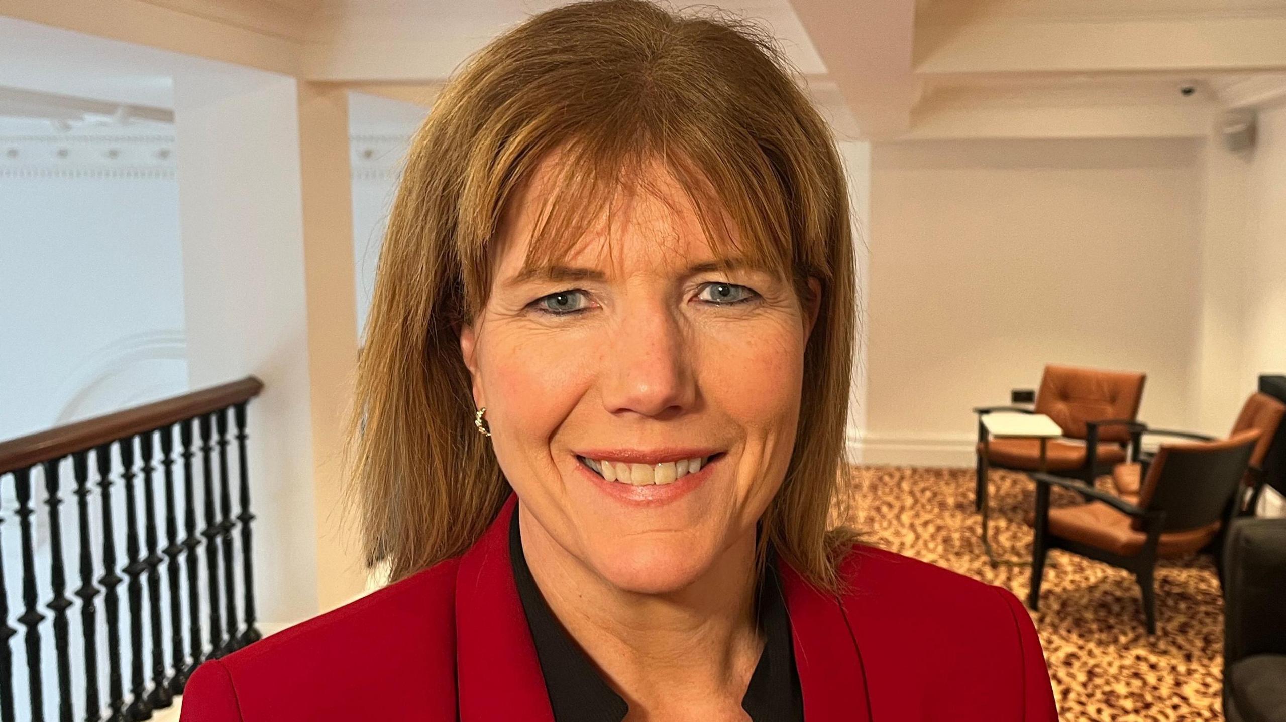 A woman with shoulder-length blonde hair is smiling at the camera. She is standing on what appears to be an indoor balcony. On the left is a railing looking onto a bigger room. On the right are chairs and a table for people to sit and chat. There is yellow and brown carpet on the floor and the walls and ceiling are white. She is earing a dark top under a red jacket.
