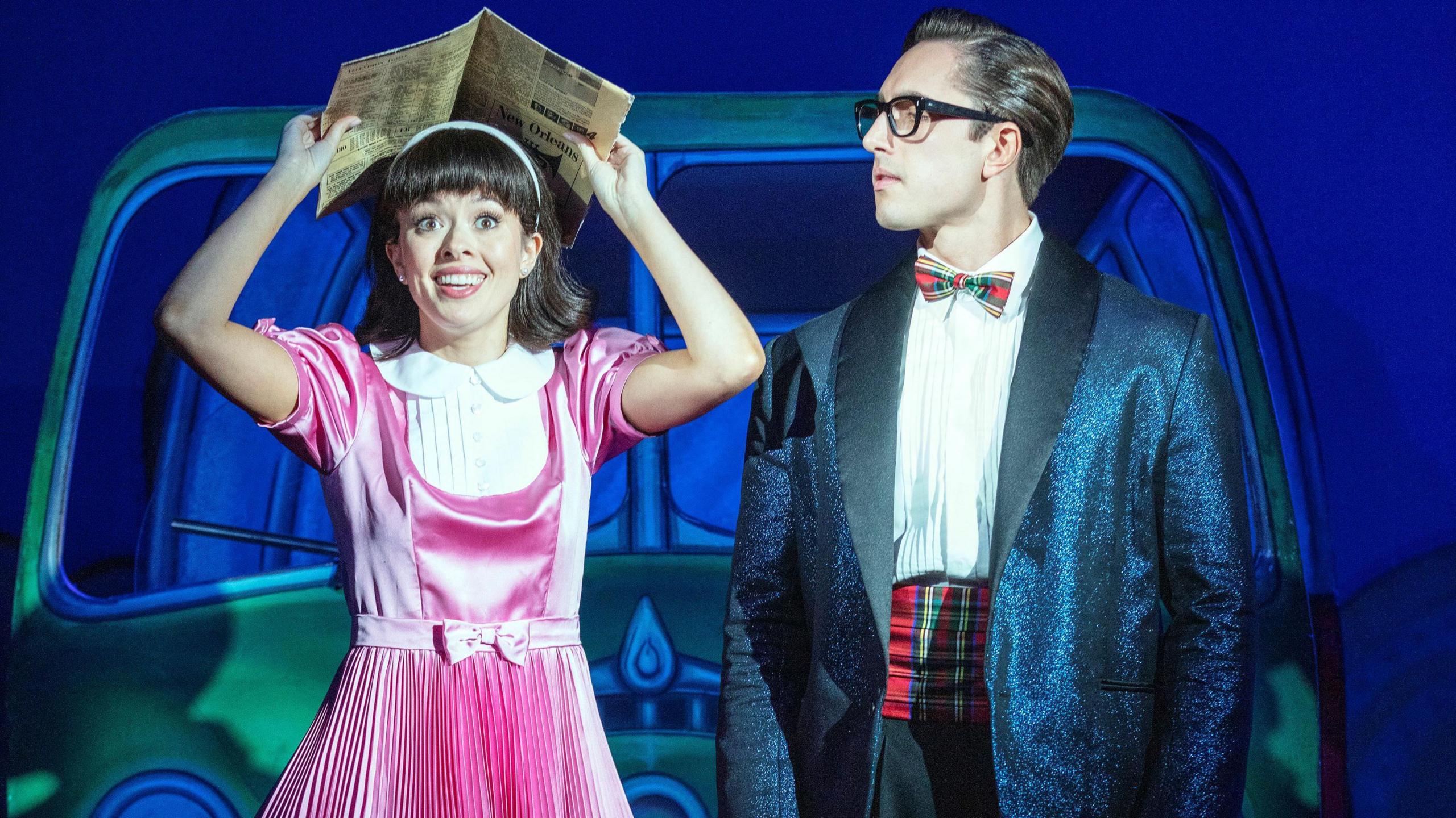 Lauren Chia is playing Janet in a pink dress, she is holding a newspaper above her head. Connor Carson is playing Brad, he is wearing a blazer, a bow tie and thick black glasses. 