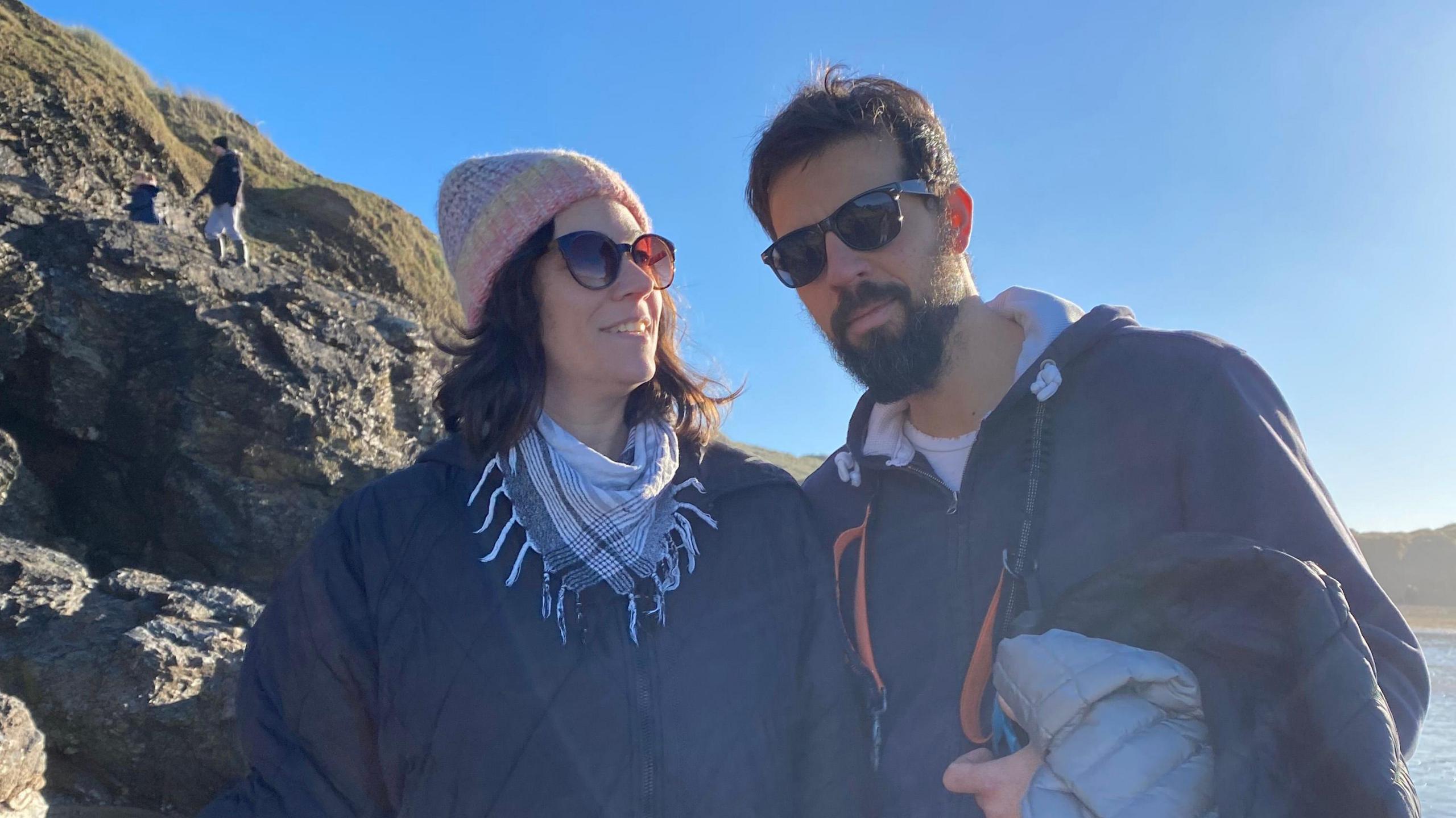 Karina Busquets, left, is wearing a coat, scarf, hat and coat, and is stood on a beah with her husband. There are rocks to the left of her and see in the background