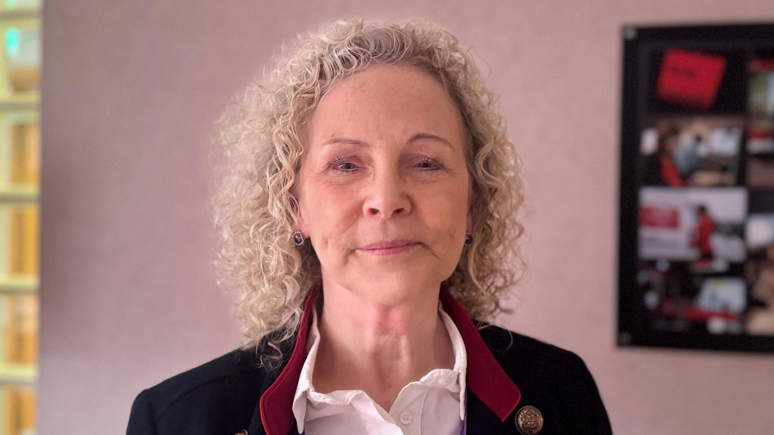 Dr Jacqui Montgomery-Devlin. She is an older woman with curly, blonde hair. She is looking at the camera with a neutral expression on her face. She is stood in a room with a pale pink wall, and a framed photo. She is wearing a white shirt, with a black blazer and red collar.