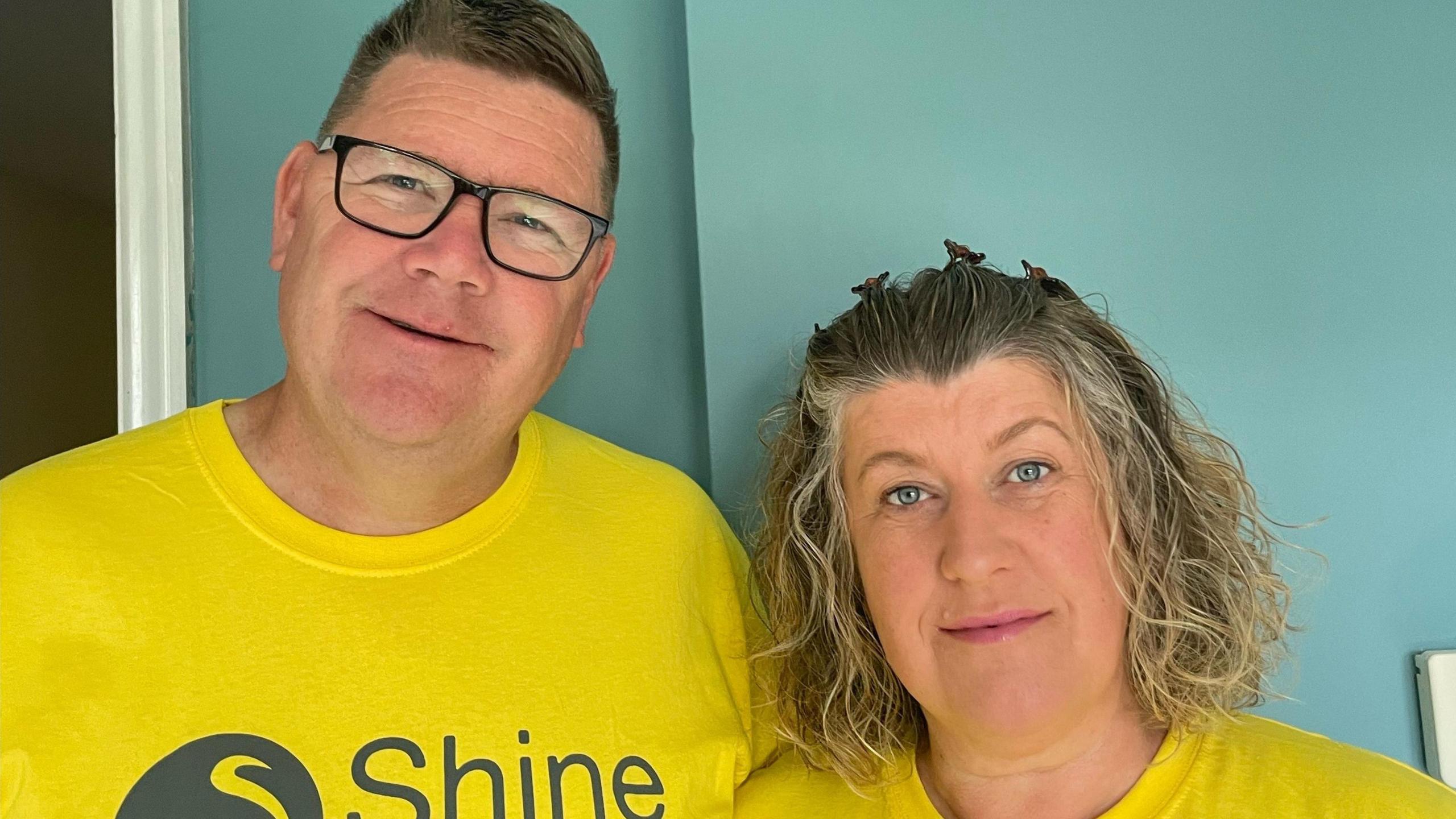 Steve and Charlotte Joynes. Steve is in the left, with brown hair and black framed glasses. His head is tilted and he is looking directly at the camera and smiling. Charlotte is stood need to him. She has short light hair and blue eyes. She is looking directly at the camera and smiling. Both of them are wearing bright yellow t-shirts with Shine written on.