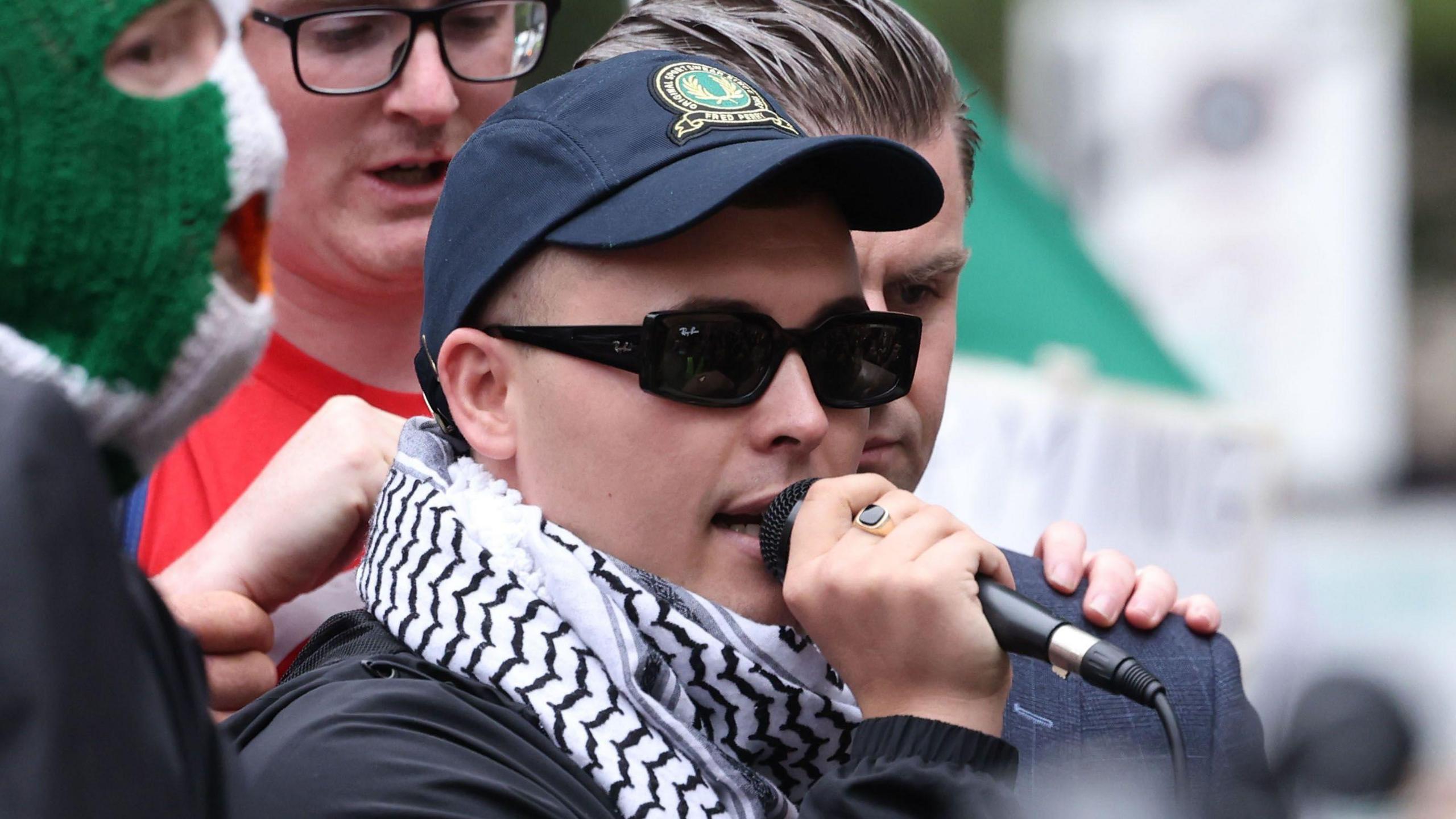 A man is photographed speaking into a microphone. He is wearing a navy blue baseball cap, dark sunglasses, a dark long-sleeve jacket and a black and white scarf.