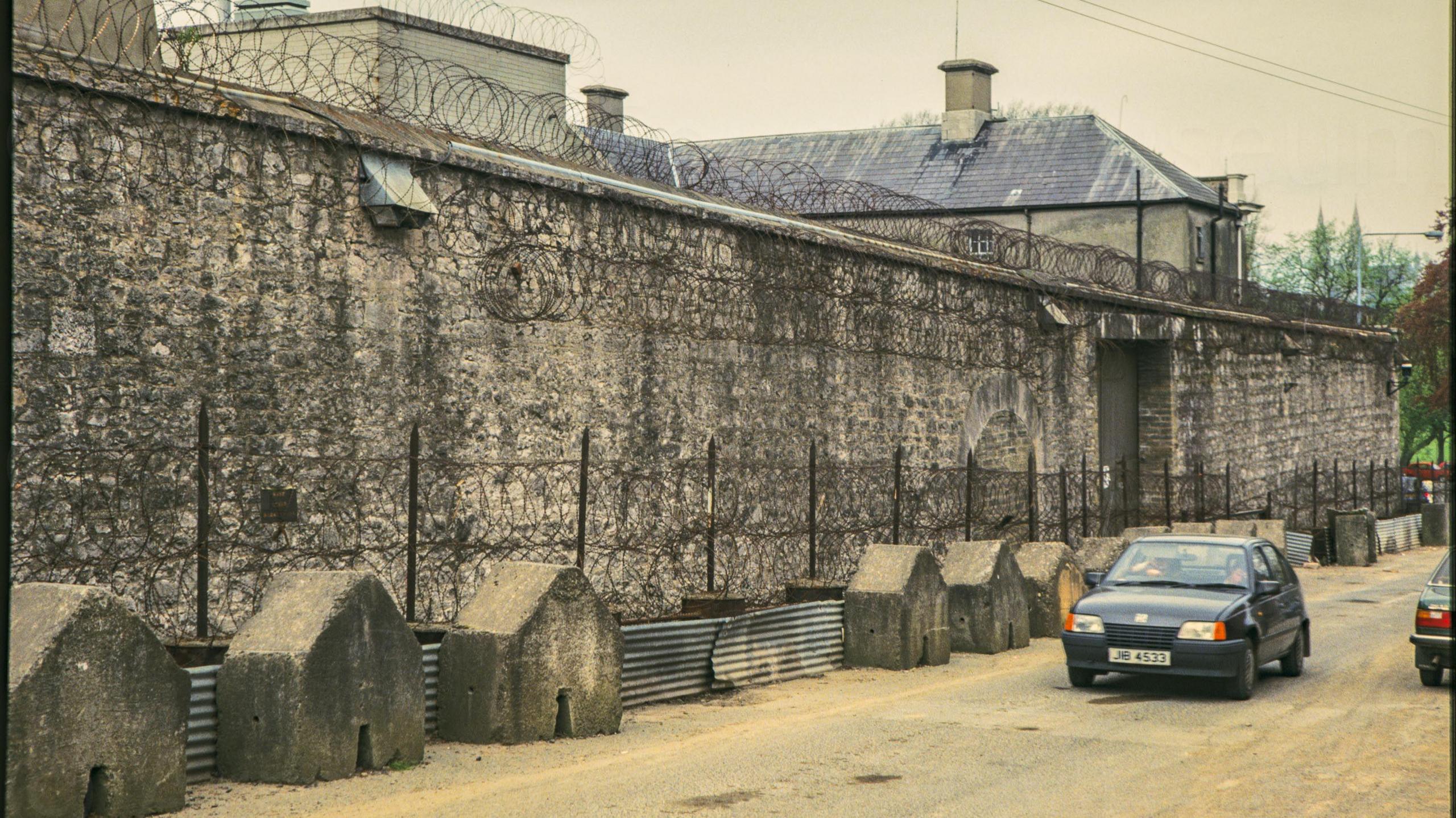 A digitalised photo from a film of a stone wall with barbed wire along the top. Looking north towards the mall showing the prison wall fortified with large concrete blocks and coils of barbed wire. The grey bricked maximum security building is partially visible over the wall.