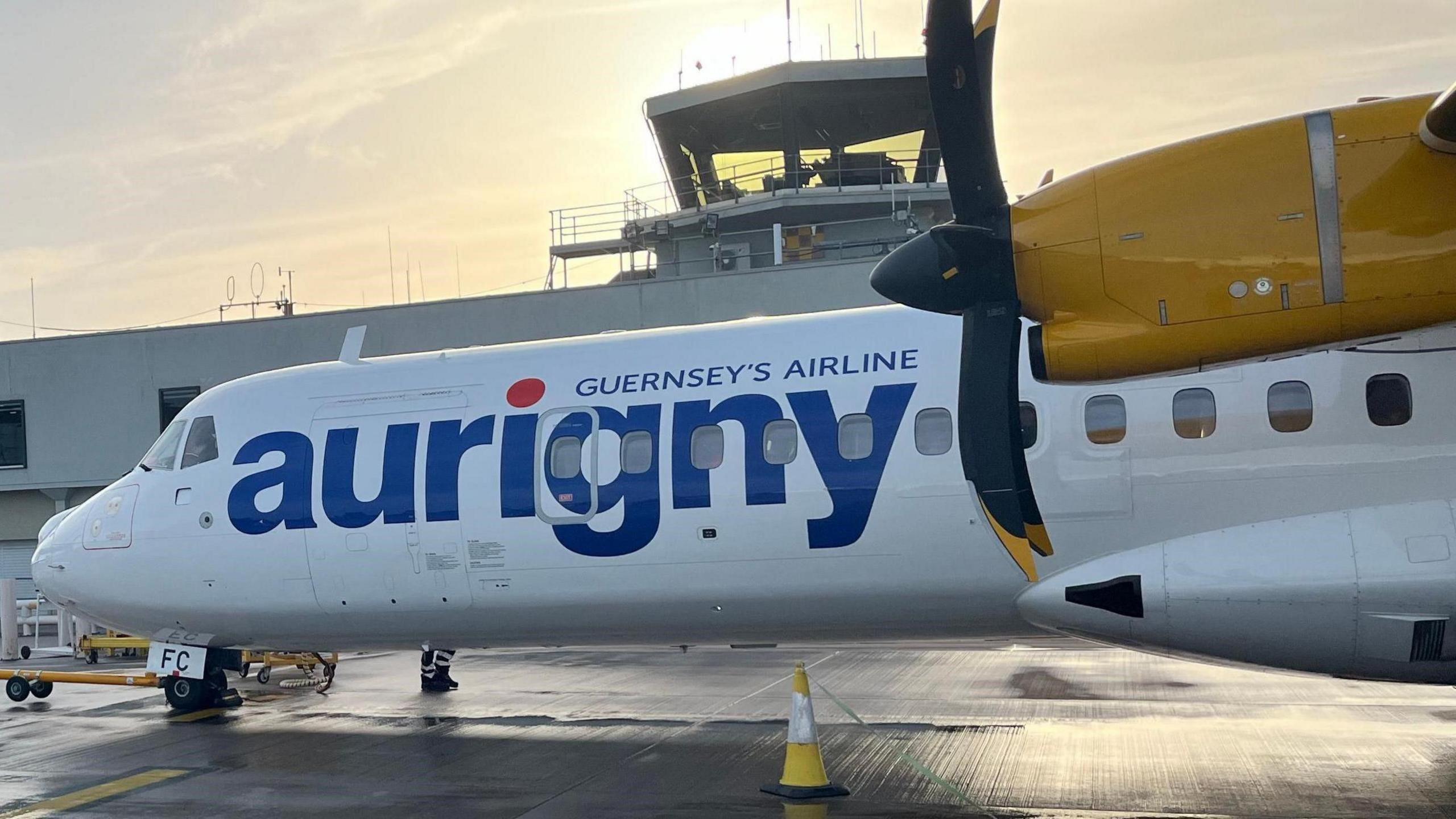 A picture of an Aurigny plane parked at Guernsey airport. It is white and yellow in colour with blue font.