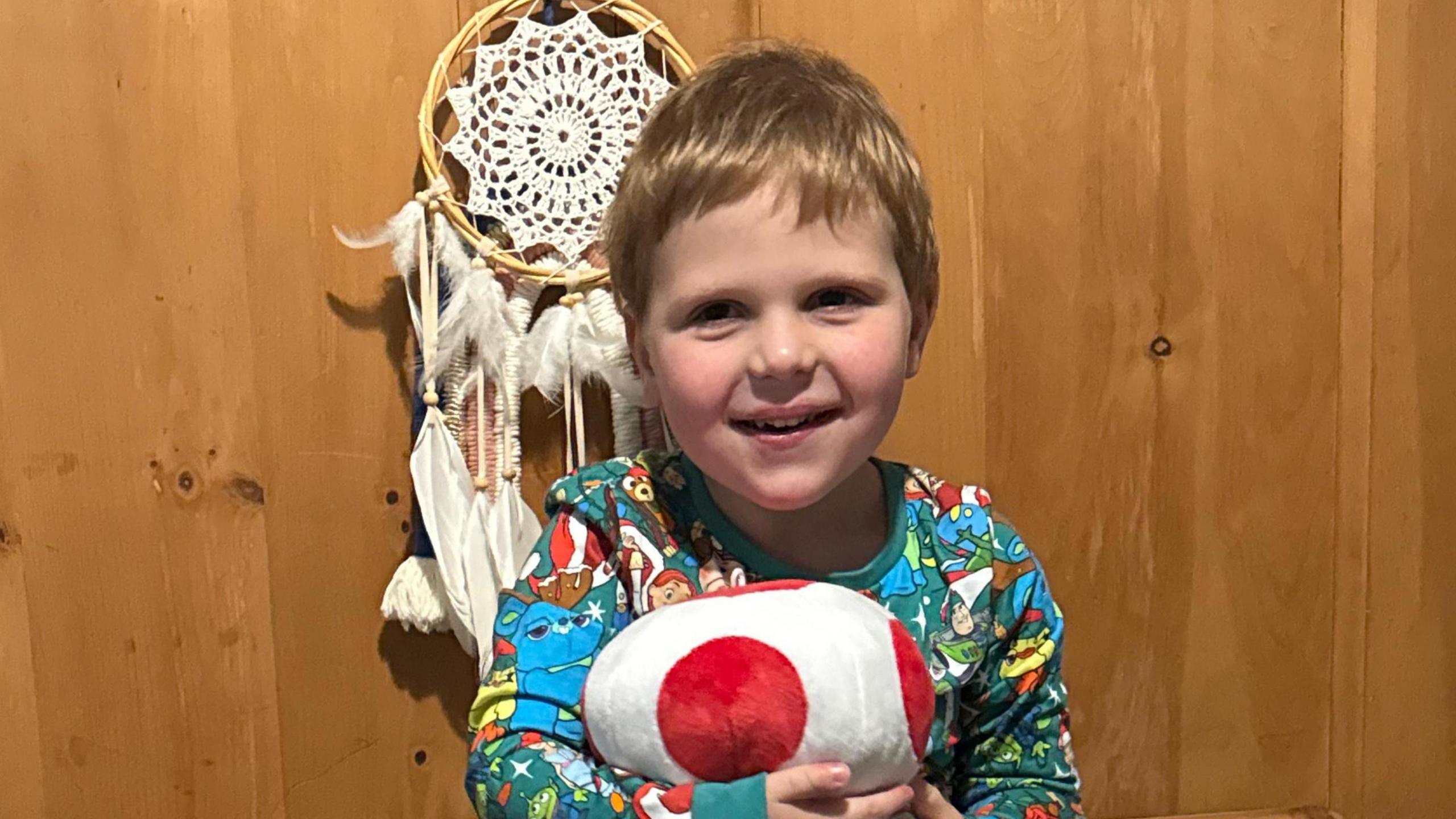 A young boy in pyjamas, holding a stuffed toy. There is a windcatcher behind him.