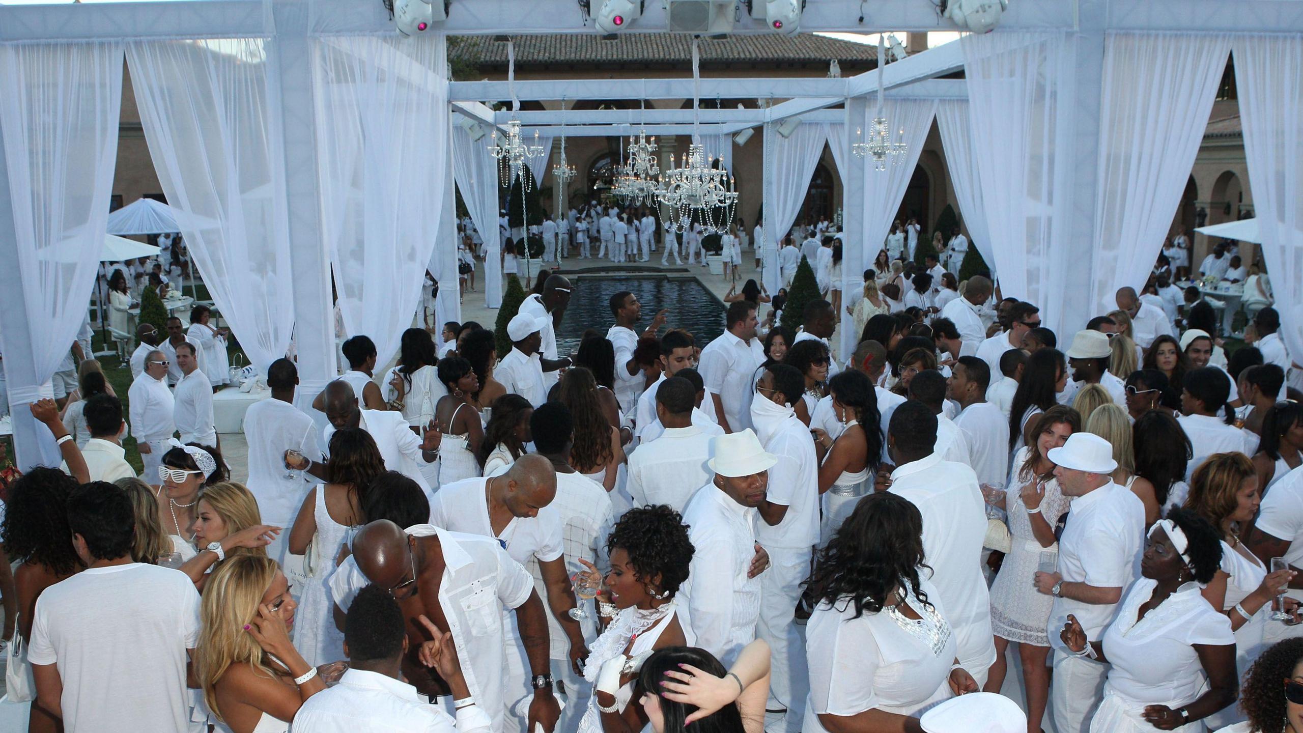 Hundreds of people - all dressed in white - huddle around a pool and tents draped with sheer white curtains. 