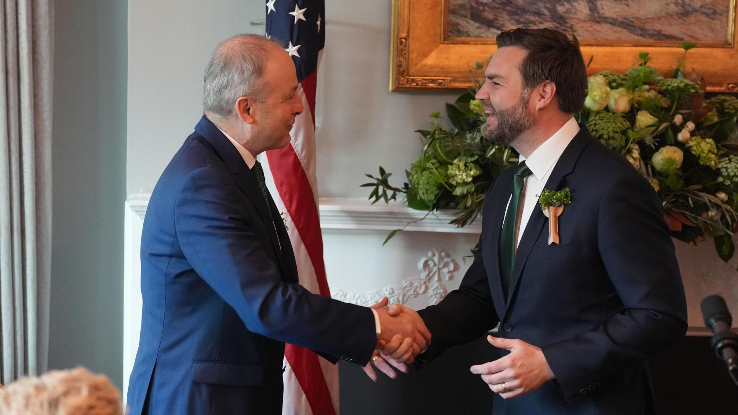 JD Vance and Micheál Martin shaking hands in front of the US flag. They are both wearing suits and smiling.